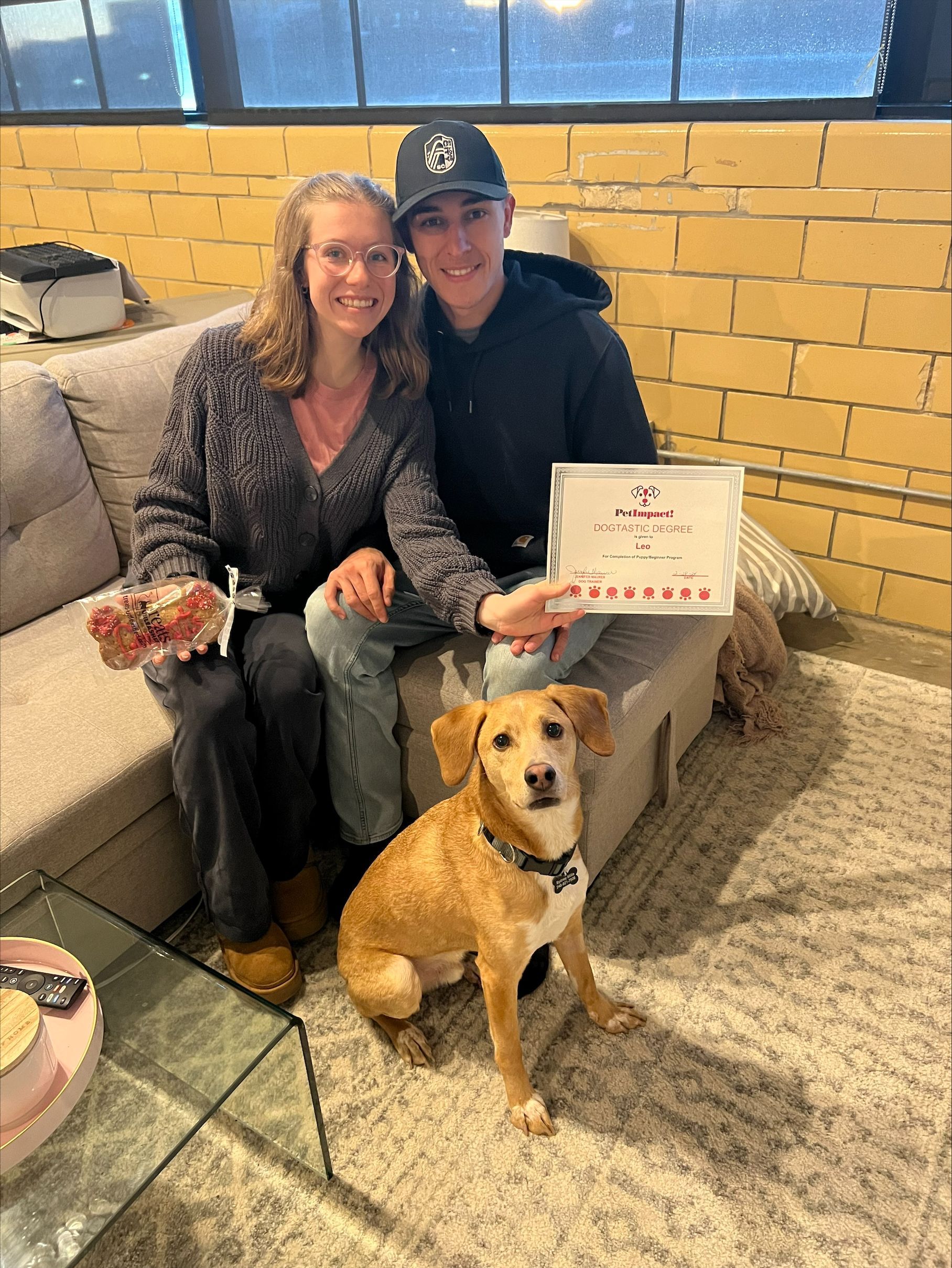 Two people seated on a couch with a dog. One holds a certificate, and the other holds a gift.  They are smiling, inside.