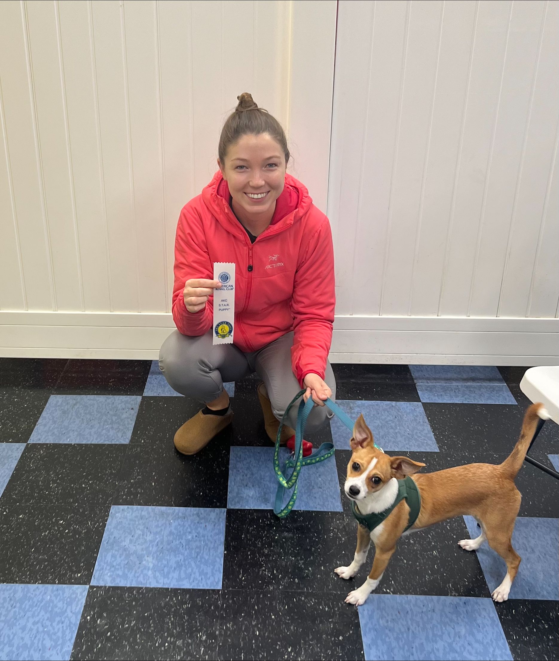 A woman is kneeling down next to a small dog on a checkered floor.