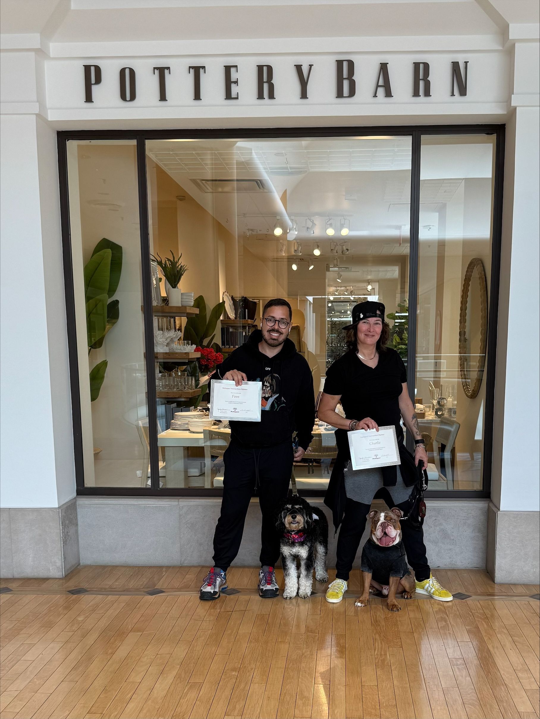 A man and a woman are standing in front of a pottery barn store with their dogs.