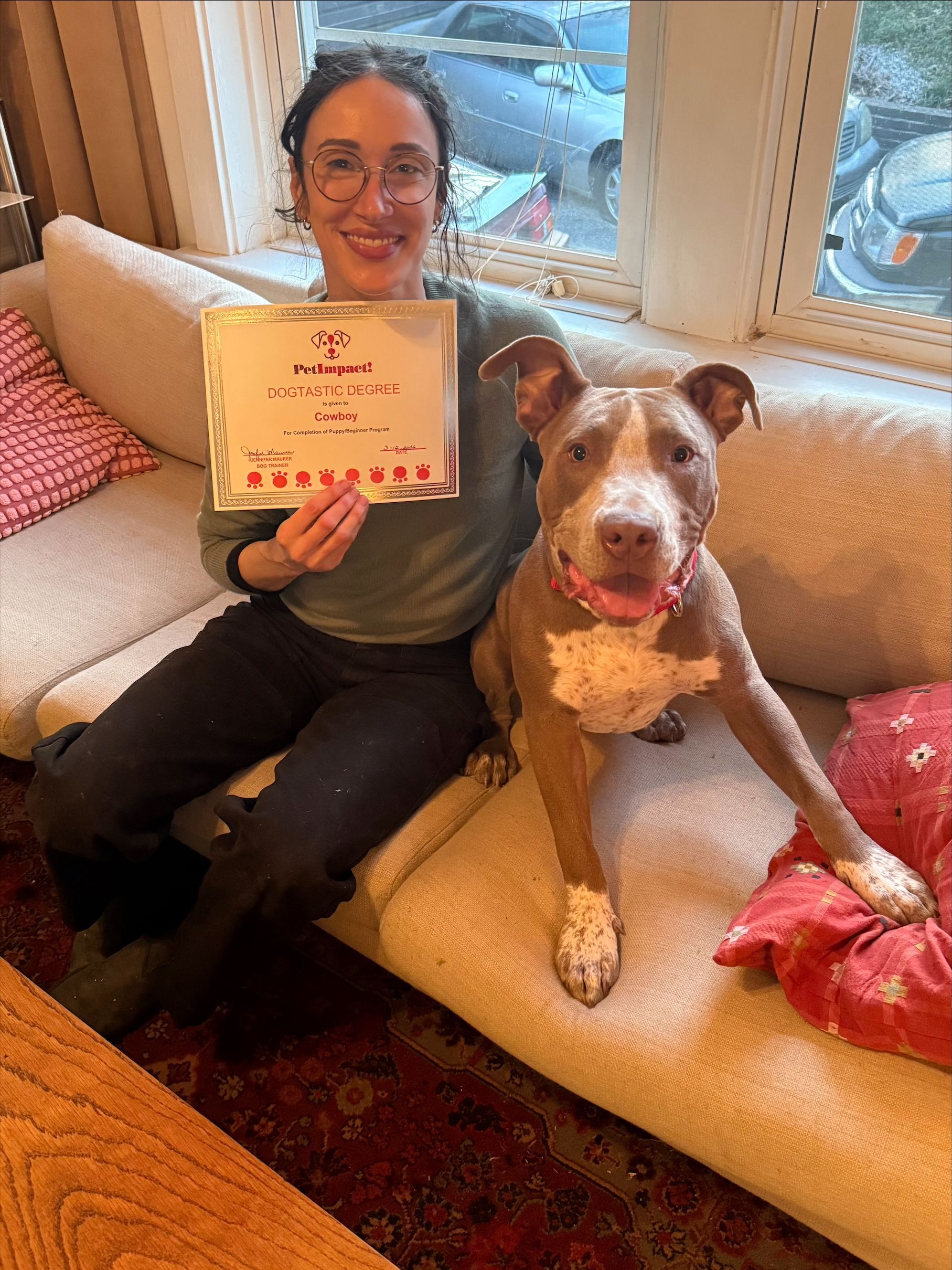 A person sits on a sofa holding a sign next to a tan and white dog that is also sitting on the couch.