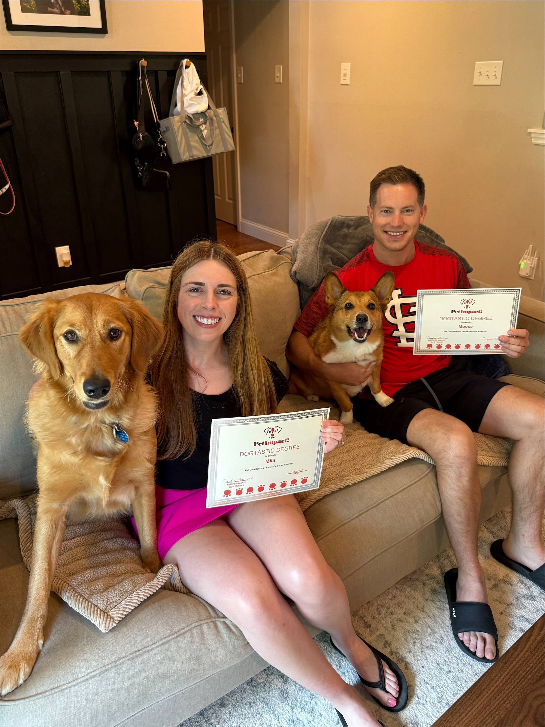 Woman and man sitting on a couch with their dogs, holding signs, smiling. Interior setting.