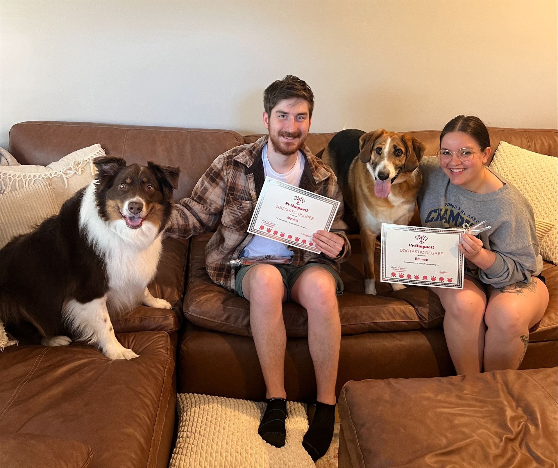 Two people sit on a brown couch with two dogs, holding certificates. One dog is brown and white, the other is black, brown, and white.