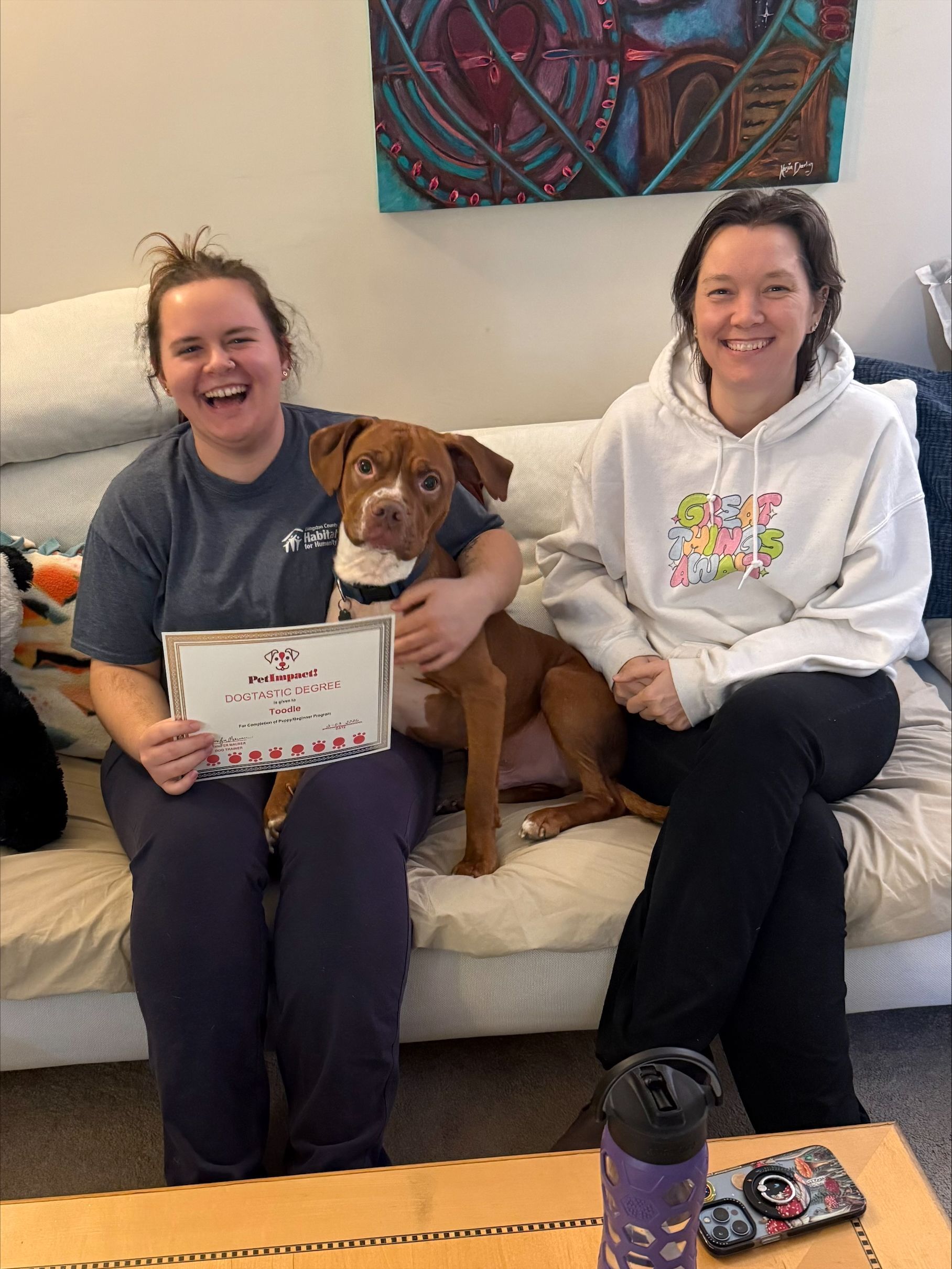 Two women and a dog on a couch. Woman on left holds a framed picture, all are smiling. Indoor setting.