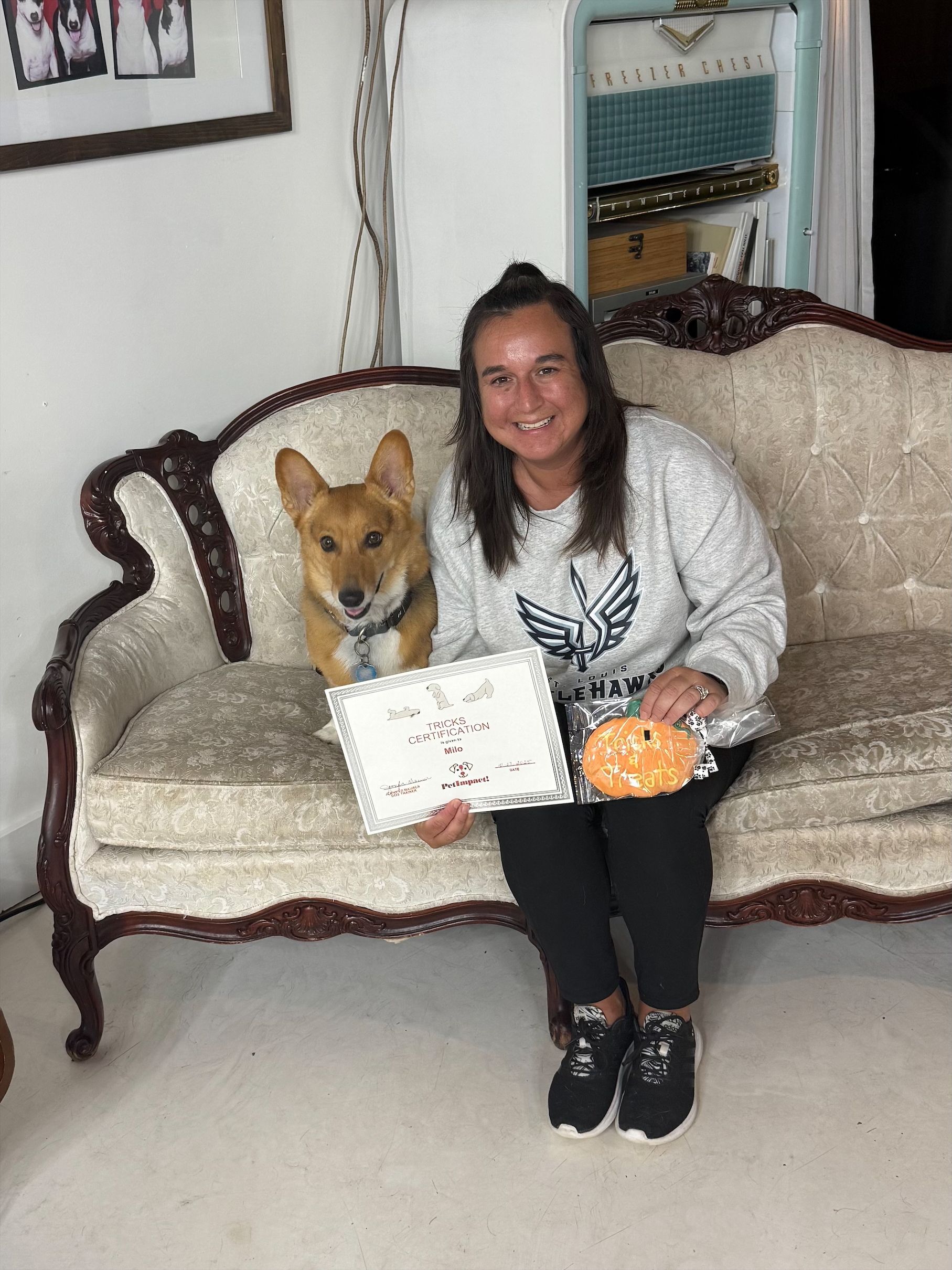 Woman and dog on ornate sofa, holding sign and pumpkin. White walls and decor in background.