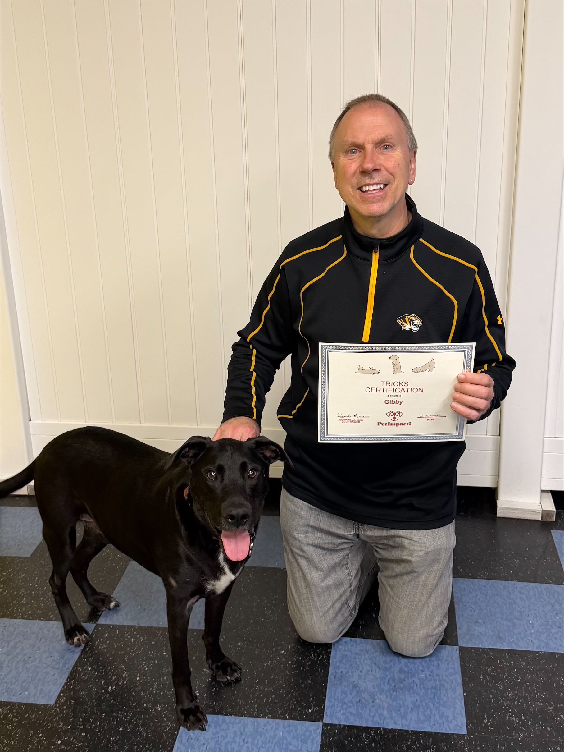 Man kneeling with a black dog, holding a certificate. They are smiling in a room with white walls and a blue floor.