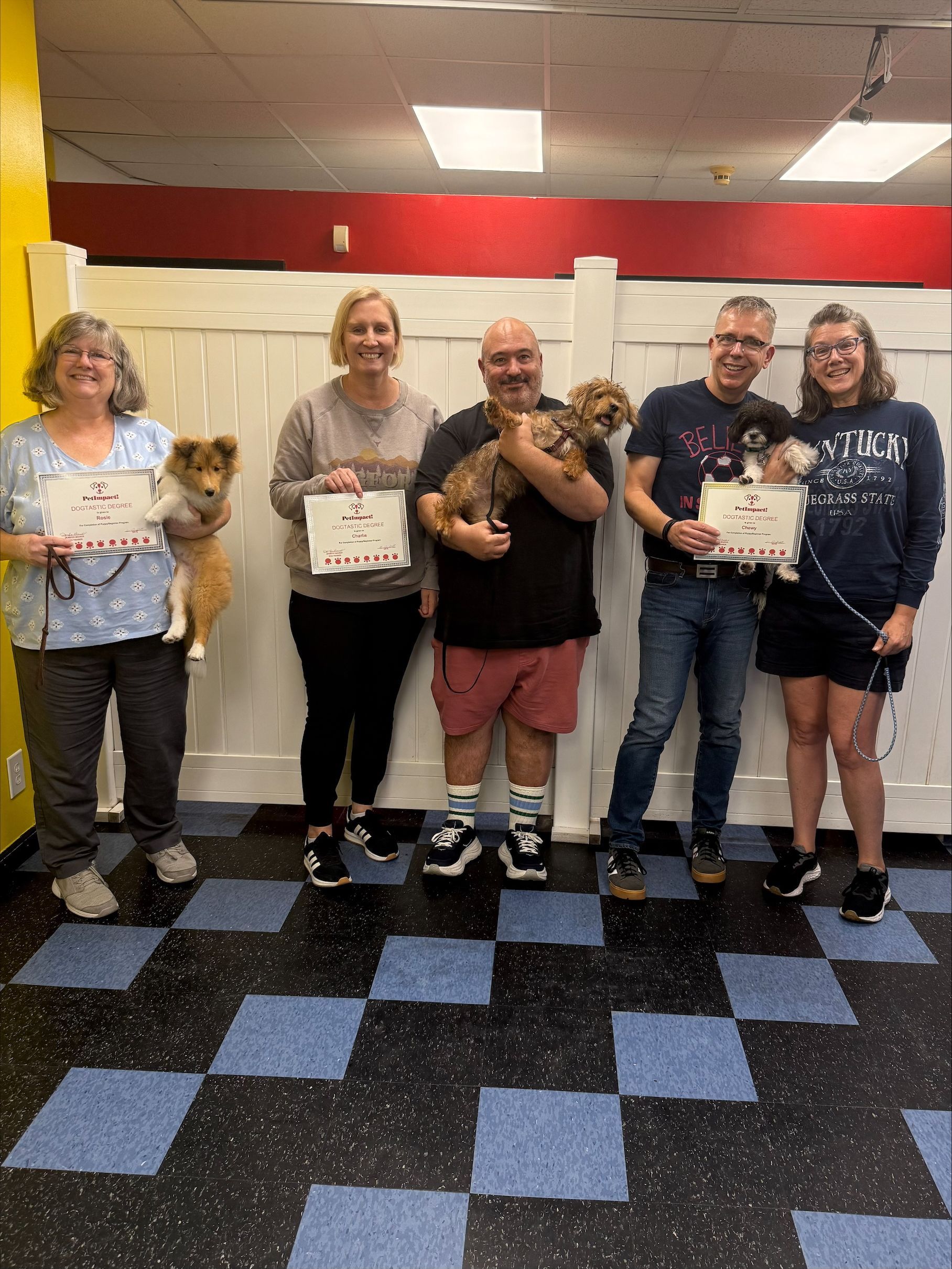 People and dogs hold certificates, posing in front of a white wall.