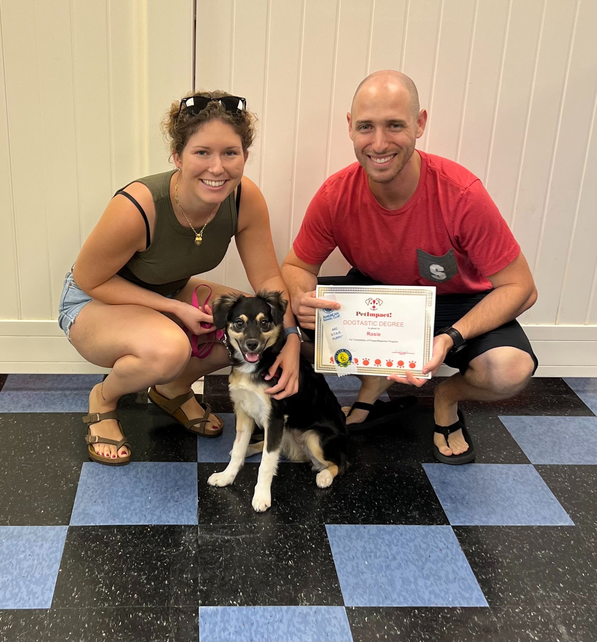 A dog sits with two people, posing with a certificate in a room with a checkered floor.