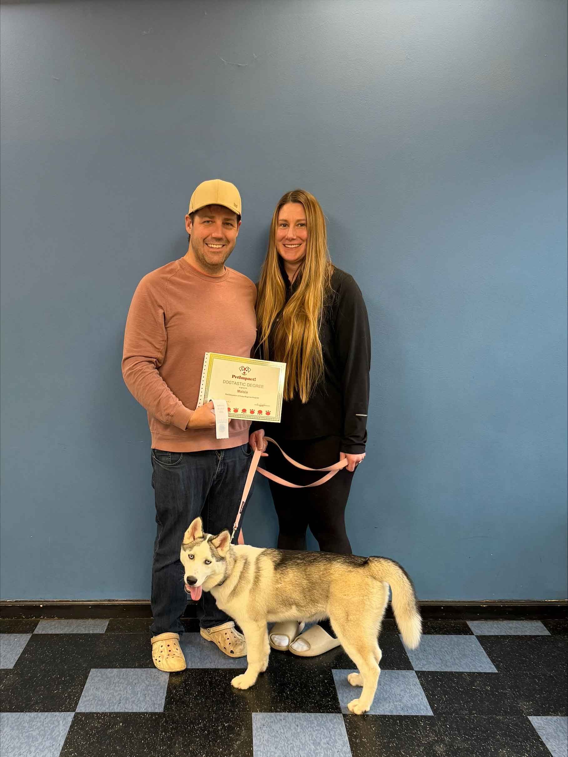A man and a woman are standing next to a husky dog on a leash.