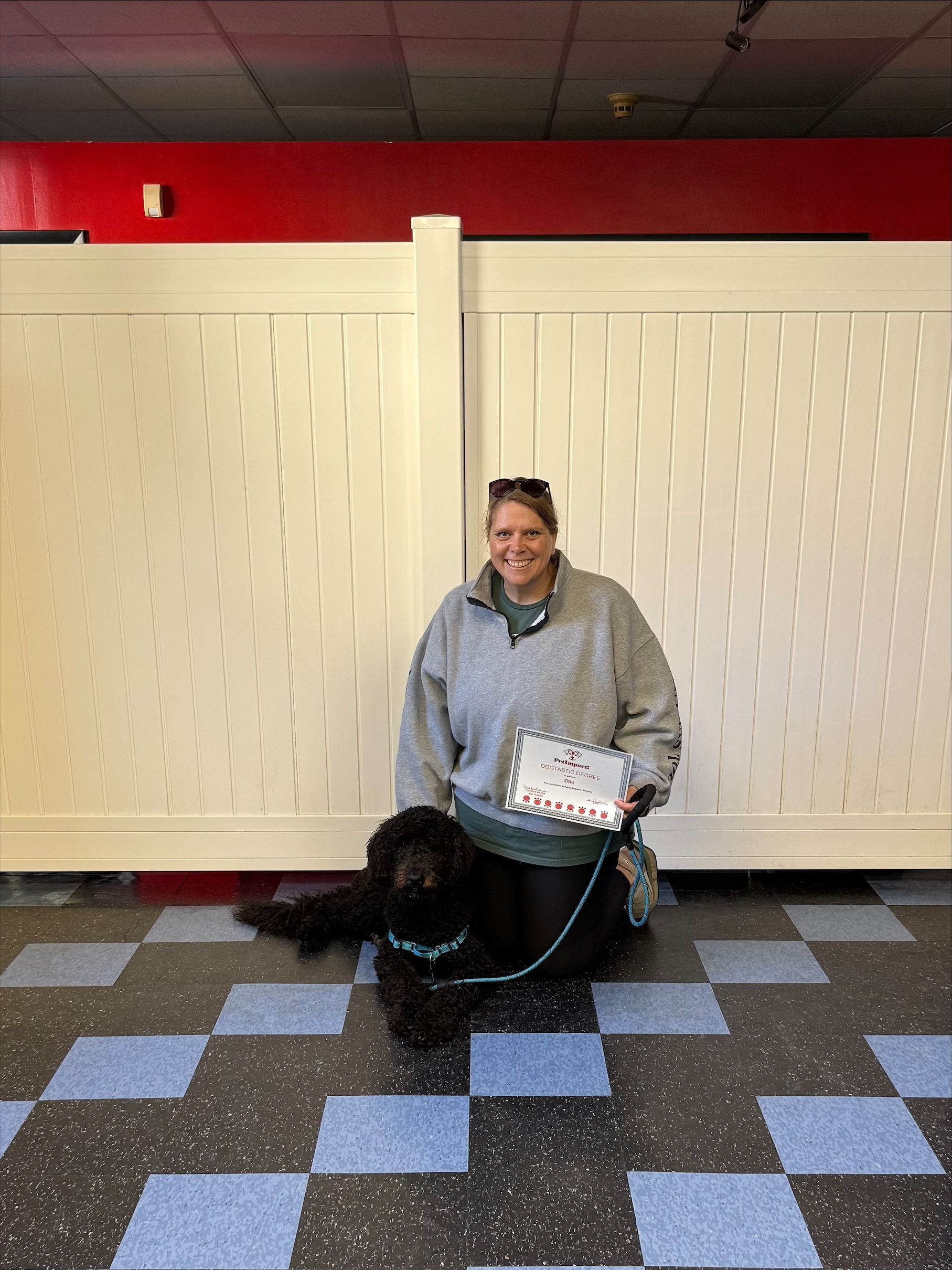 Woman kneeling with black dog; holding sign, in front of a white fence, tiled floor.