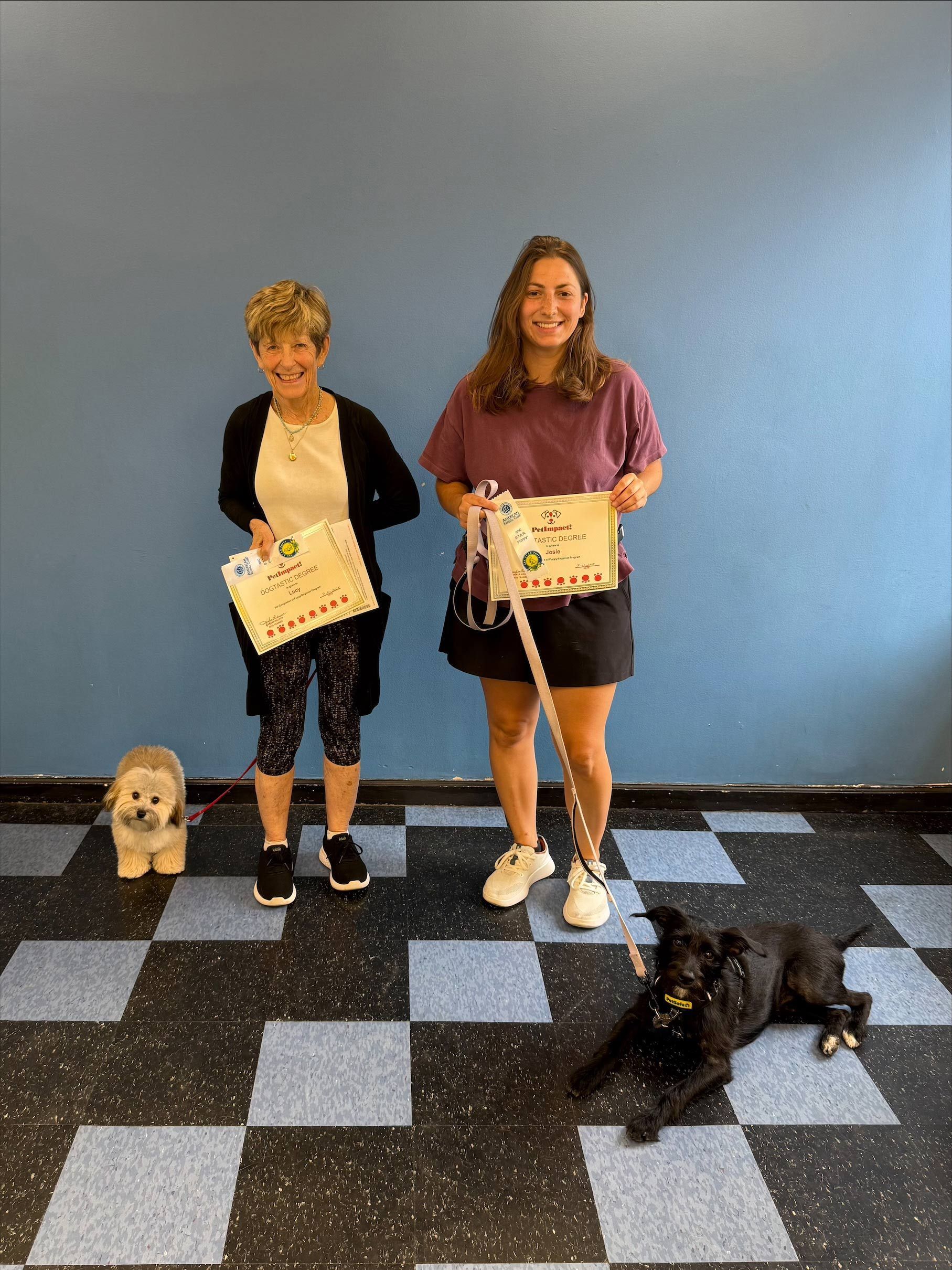 Two women hold signs, smiling, with two pets on a blue and black floor, against a blue wall.