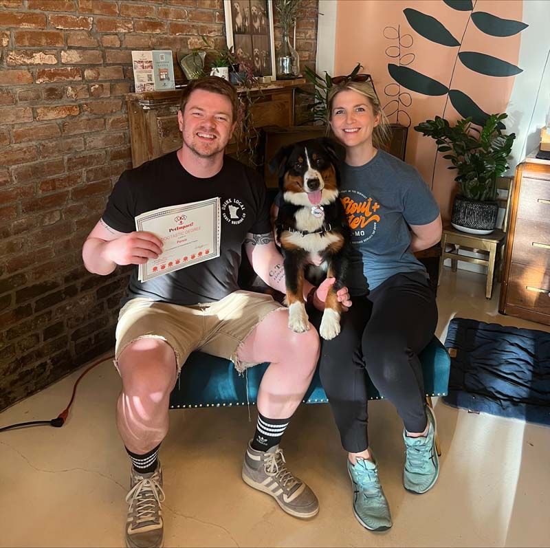 A couple with their dog sits on a blue bench, man holds a certificate. Interior with brick wall and plant.