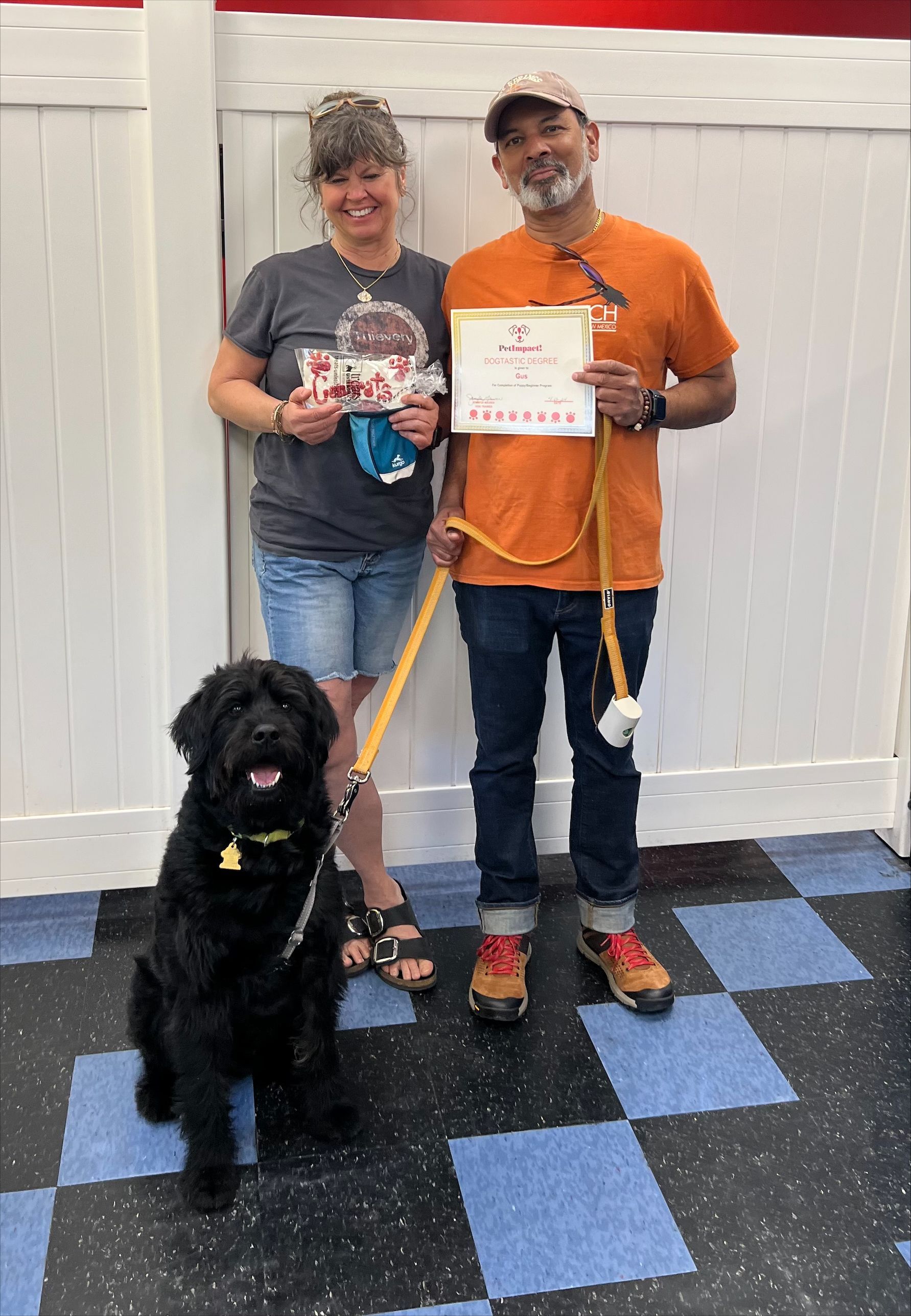 A couple and a black dog stand in front of a white wall. The couple holds awards. The dog sits.