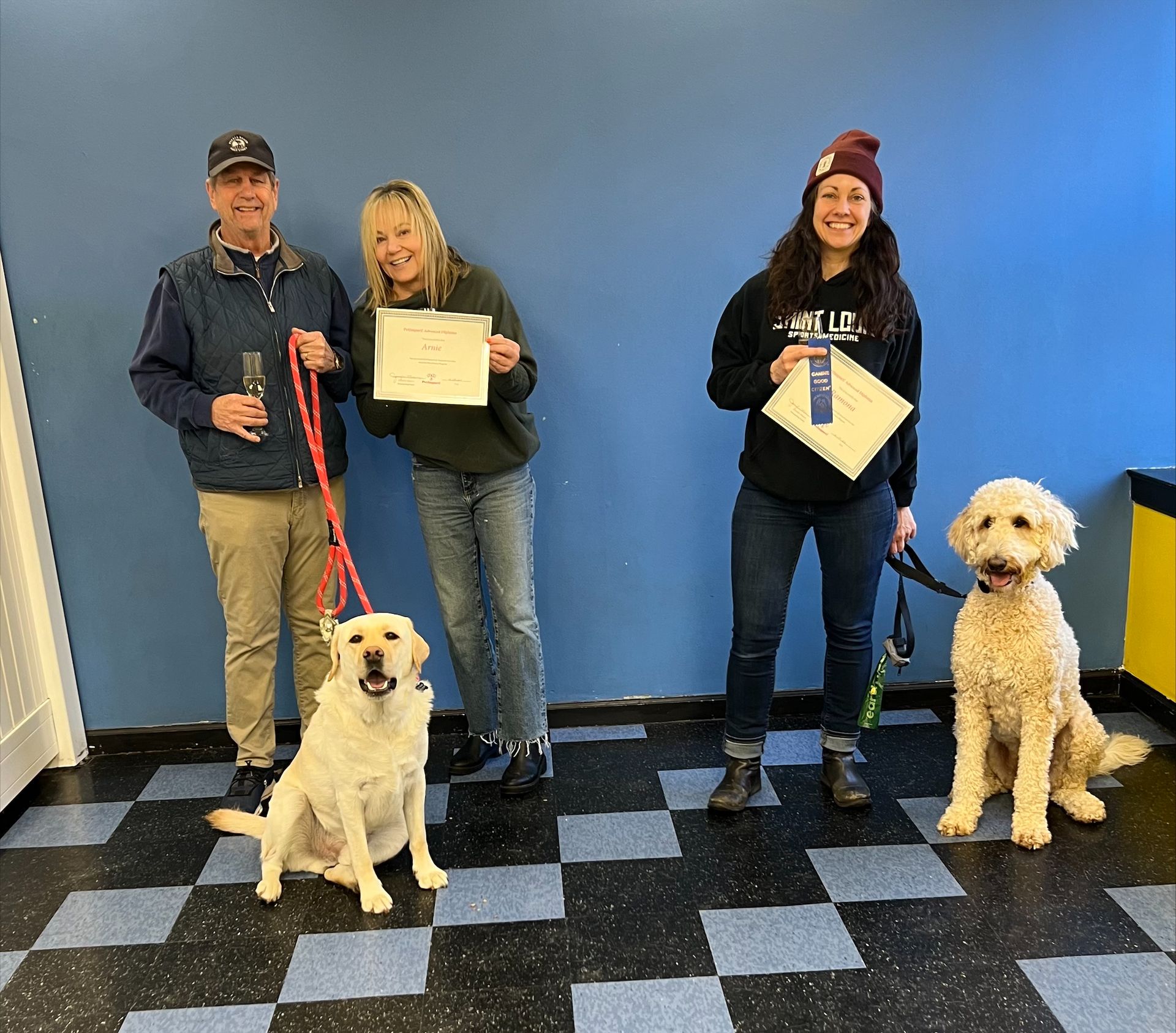 Three people and two dogs stand in front of a blue wall holding awards. A checkered floor.