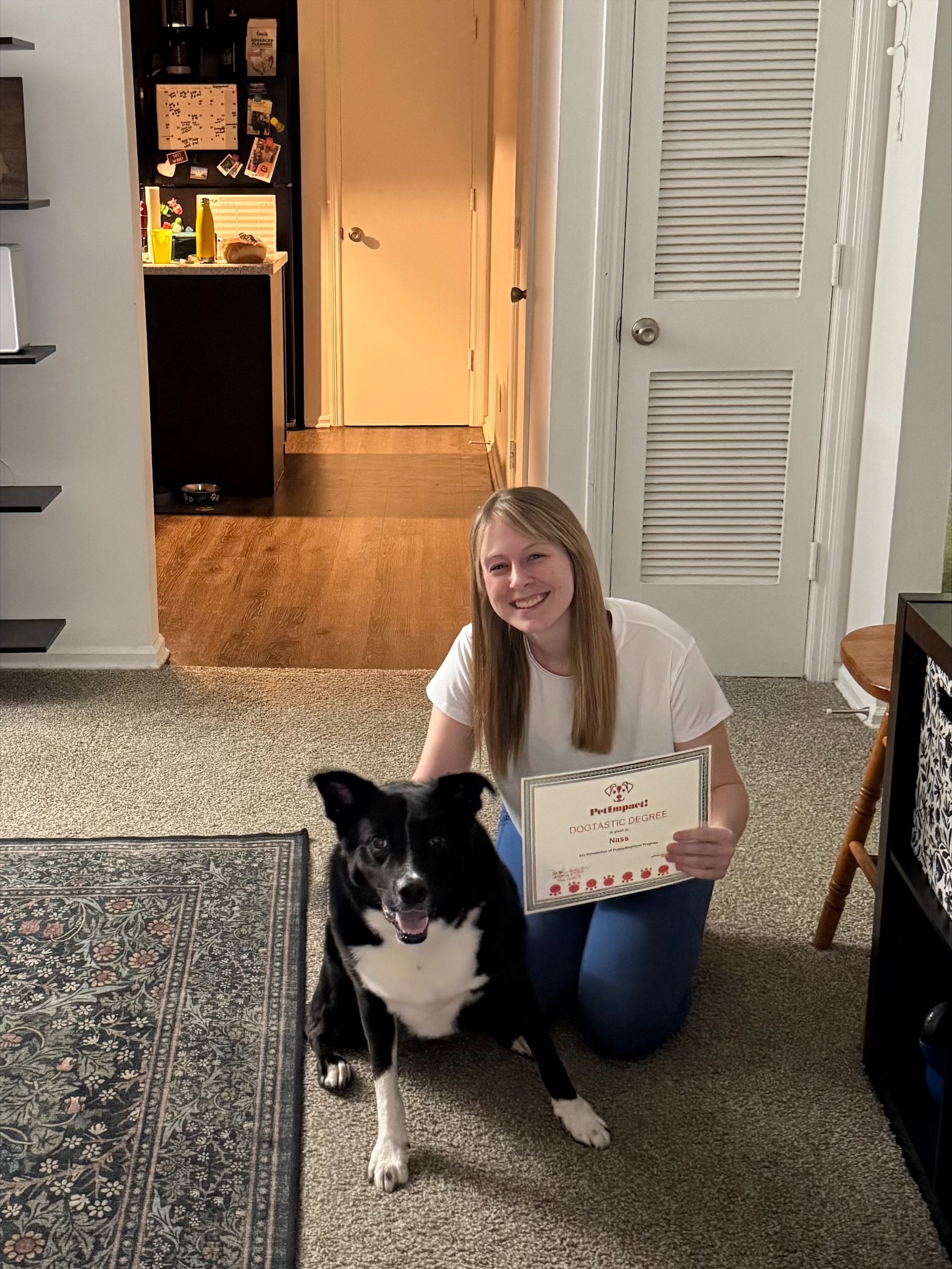 Woman kneeling next to a black and white dog, holding a certificate indoors.