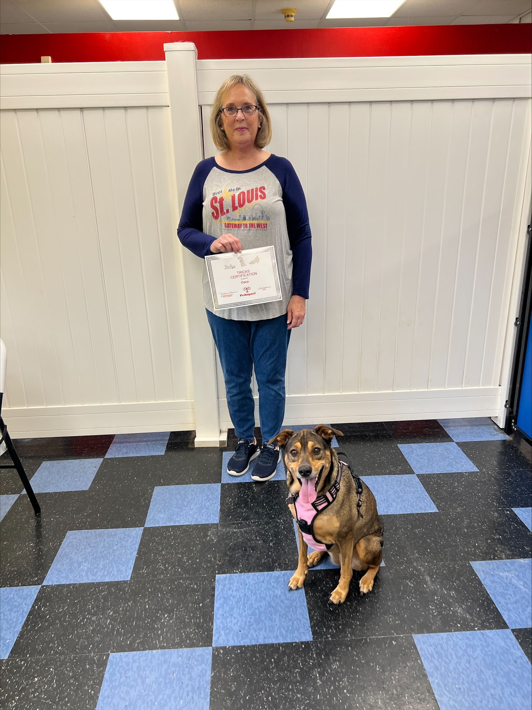 Woman and dog pose, celebrating. Woman holds a certificate, wearing a St. Louis shirt, jeans, and glasses. Blue and black checkered floor.