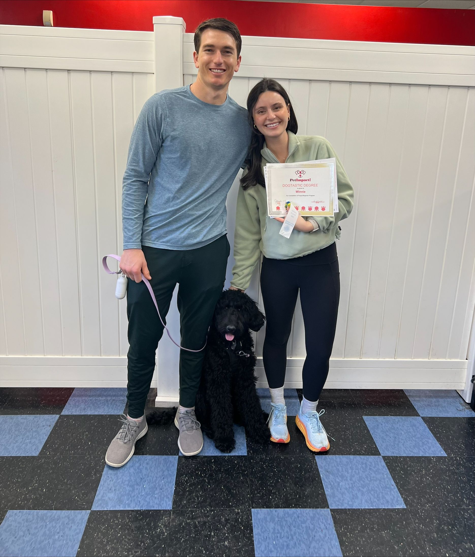 Two people and a black dog, smiling, holding a certificate. Indoors, against a white wall with red and blue flooring.