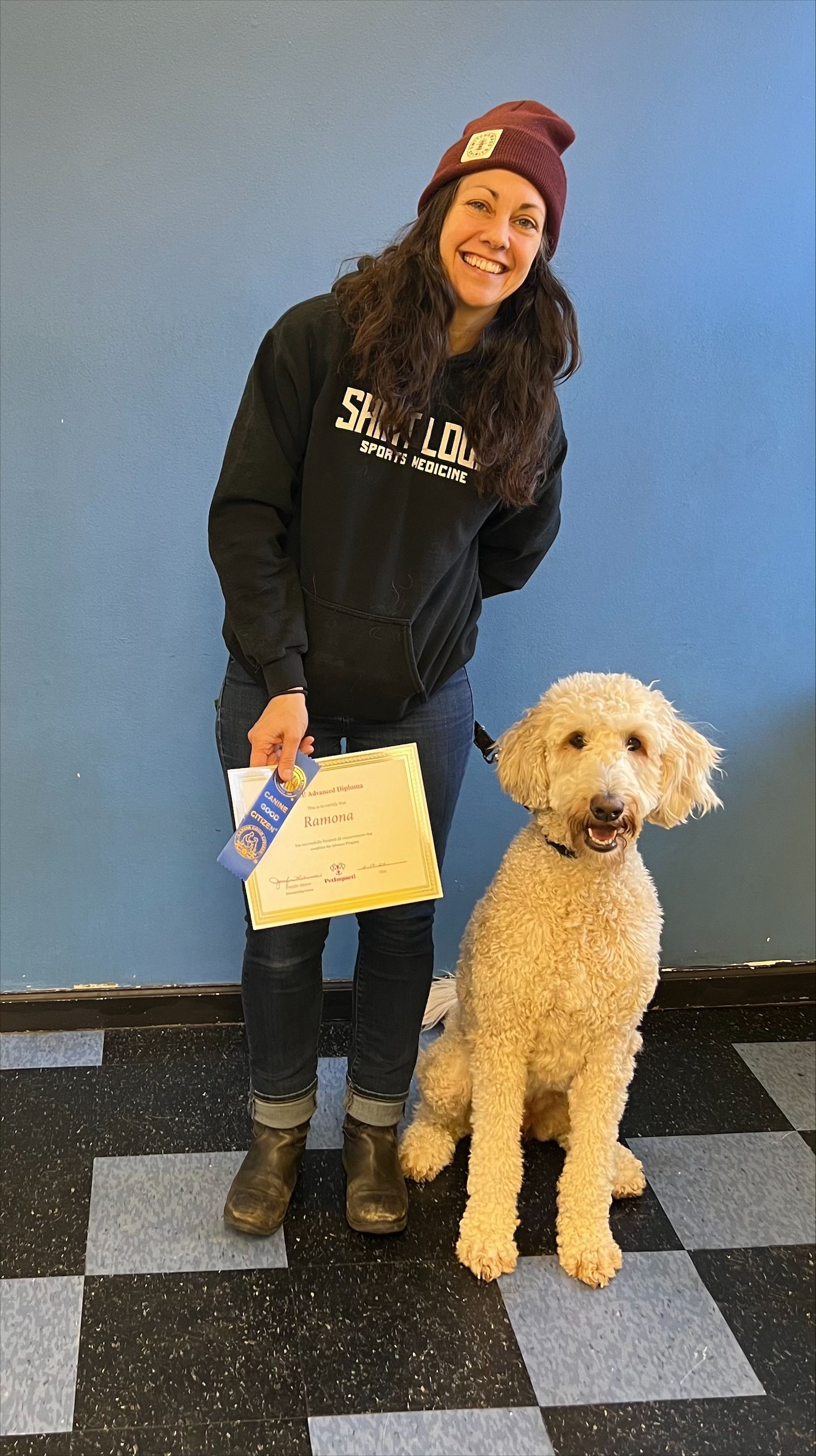 Woman in a beanie and sweatshirt, with a tan poodle. They stand in front of a blue wall and checkered floor.