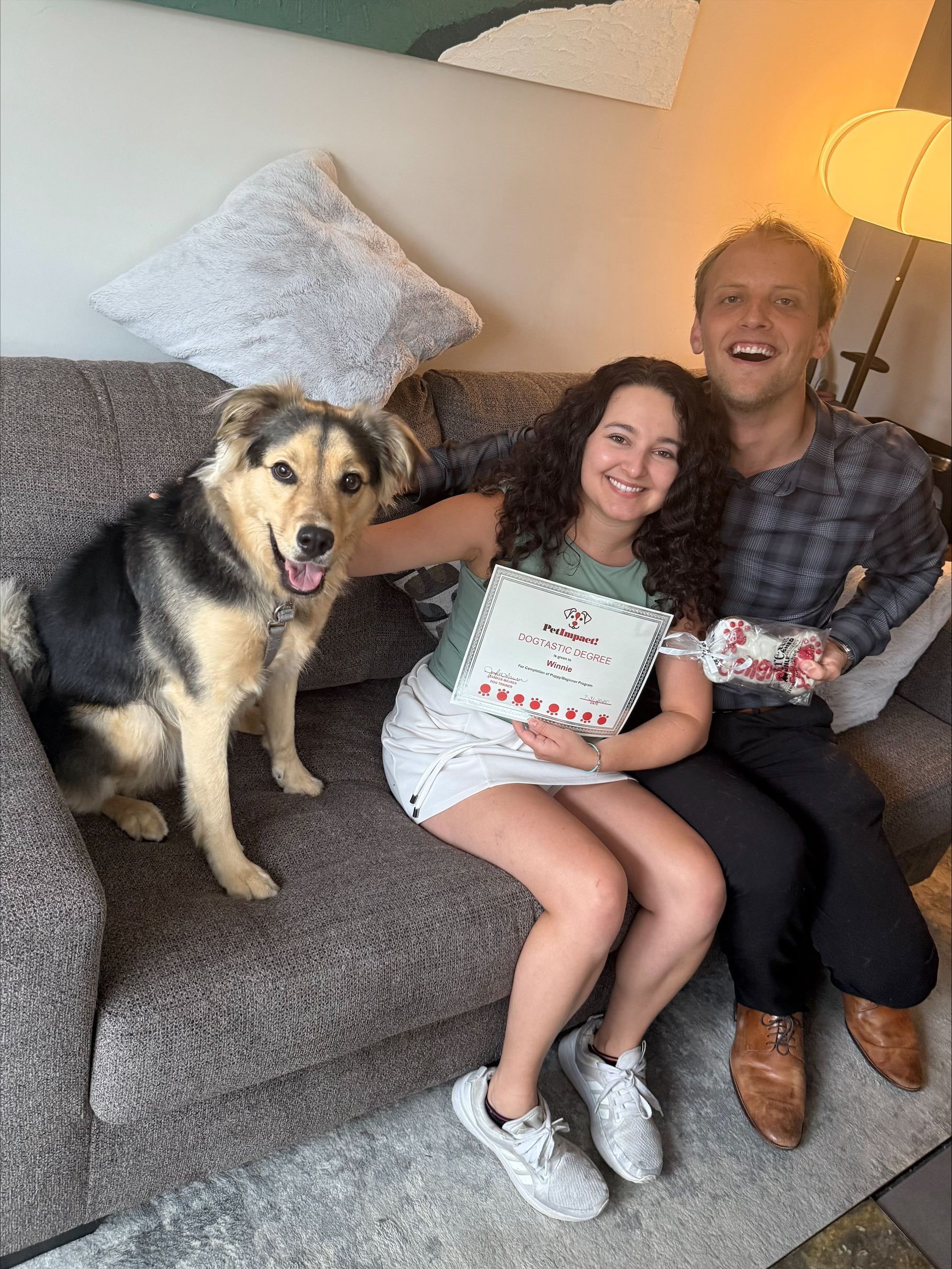 A couple and a dog sit on a couch. The woman holds a certificate, and the man is laughing. The dog looks at the camera.