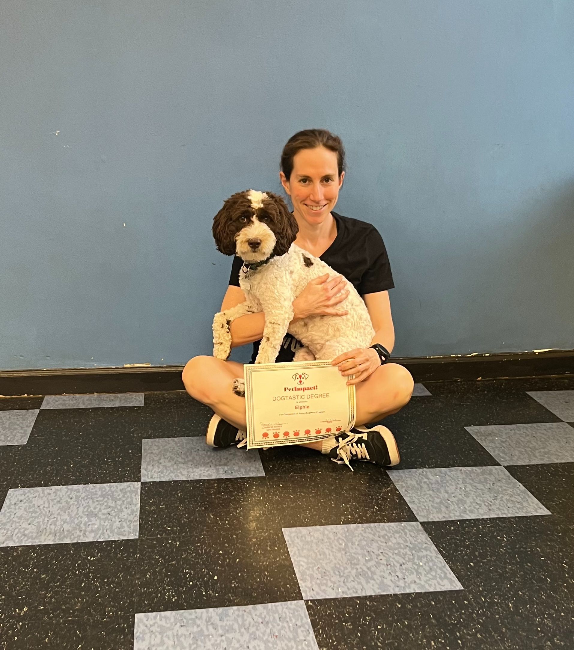 Woman seated cross-legged, holding a dog and a certificate against a blue wall and checkered floor.