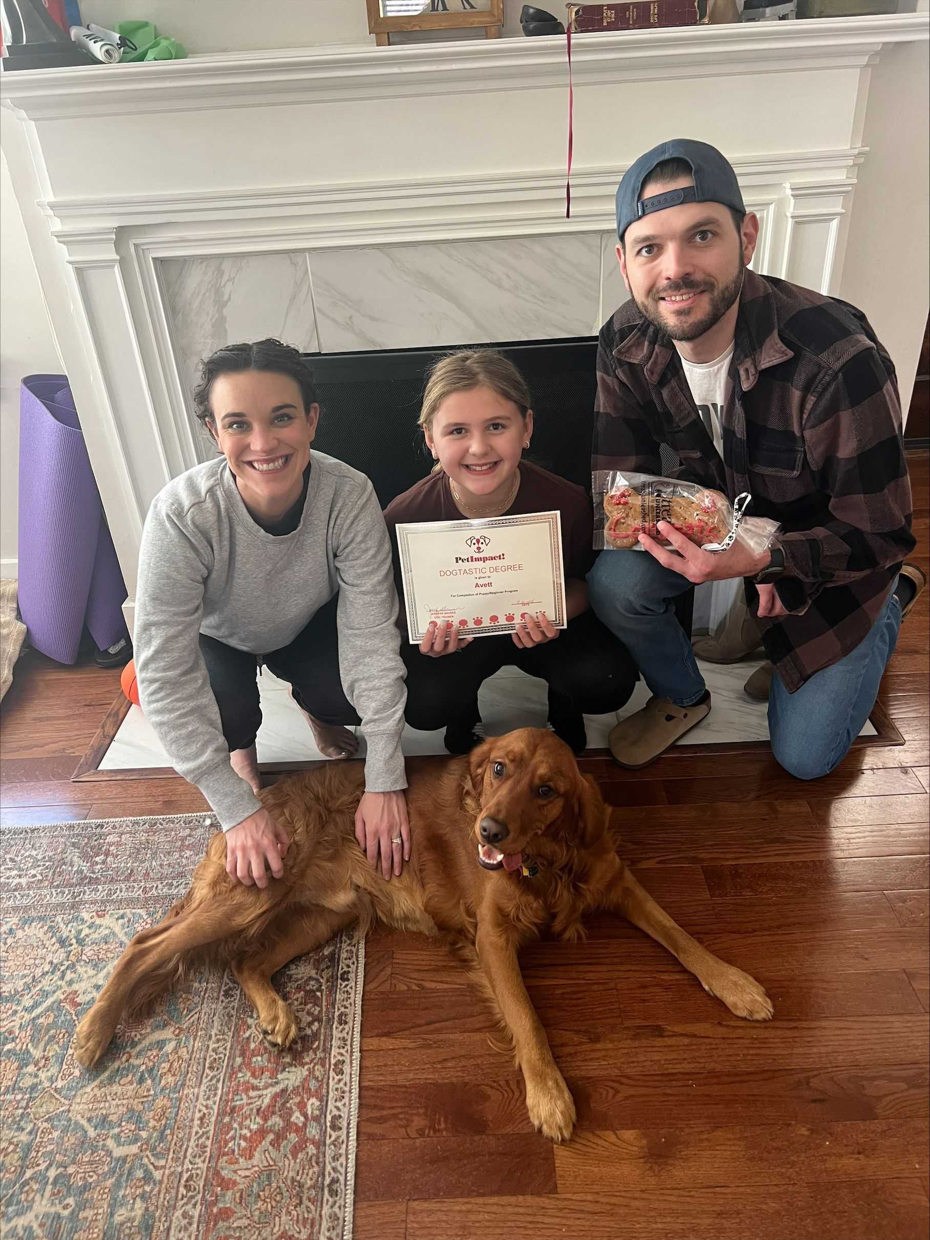 Family, including a dog, kneels in front of a fireplace. Holding treats and a card, smiles.