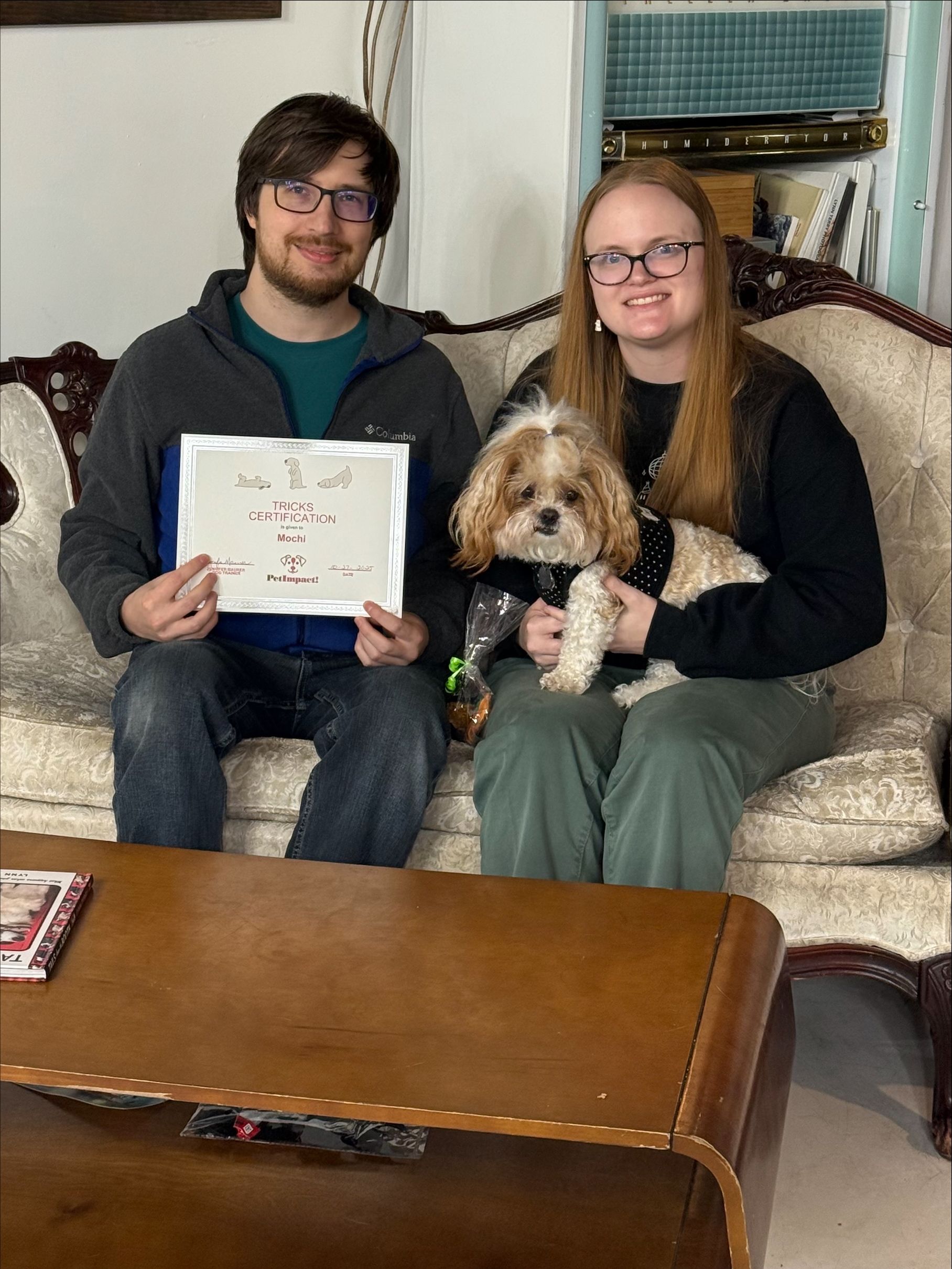 Couple with a dog on a couch, holding a certificate. They are smiling. Living room setting.