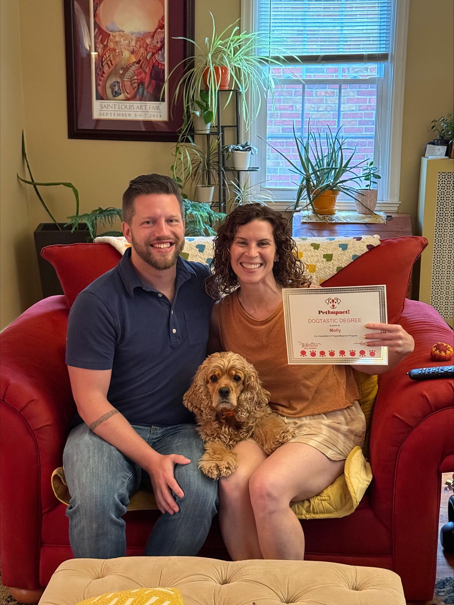 A smiling couple and their dog sit on a red couch, holding a certificate. The setting is a living room with plants and a window.