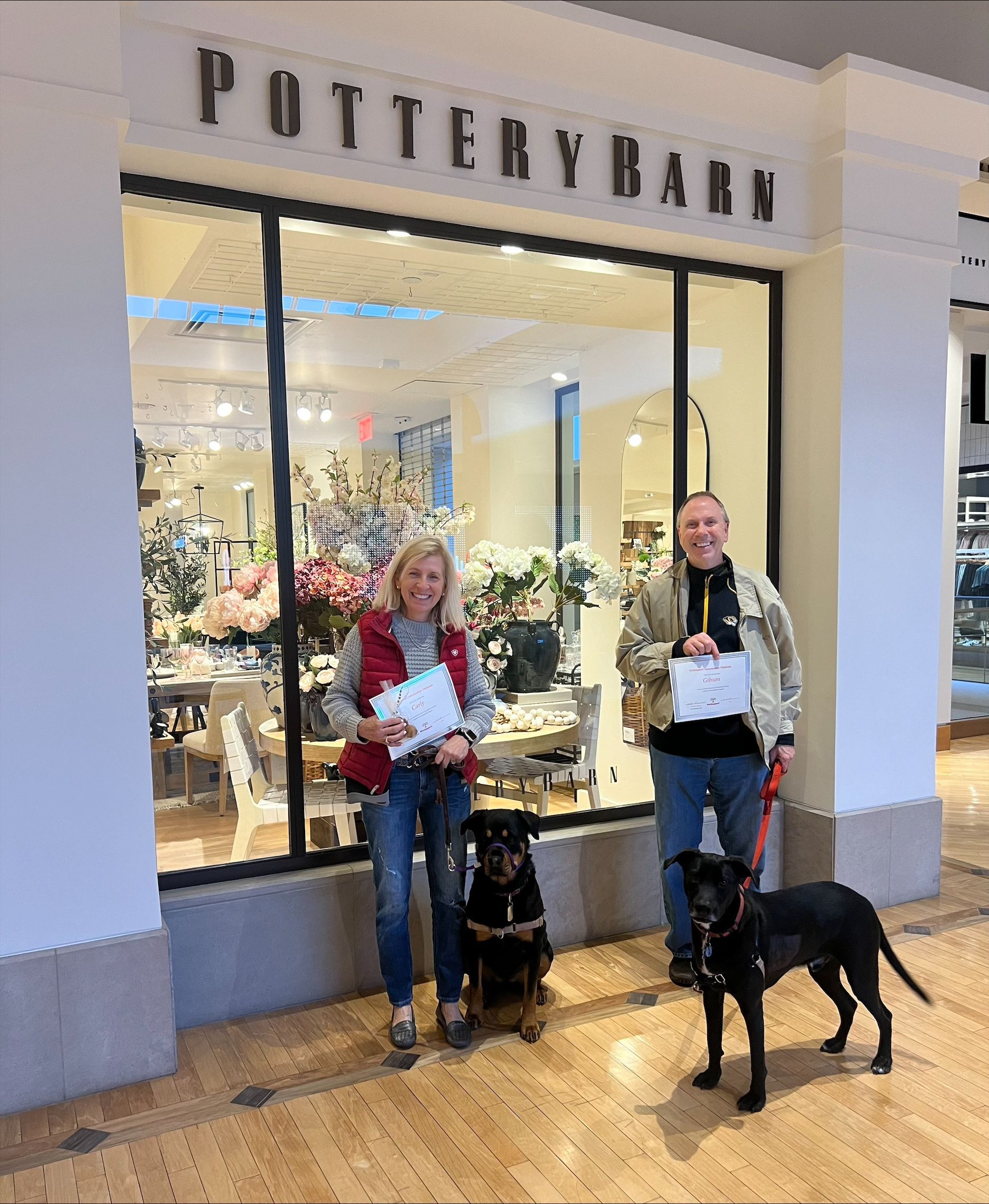 Two people with dogs stand outside a Pottery Barn store, holding papers.