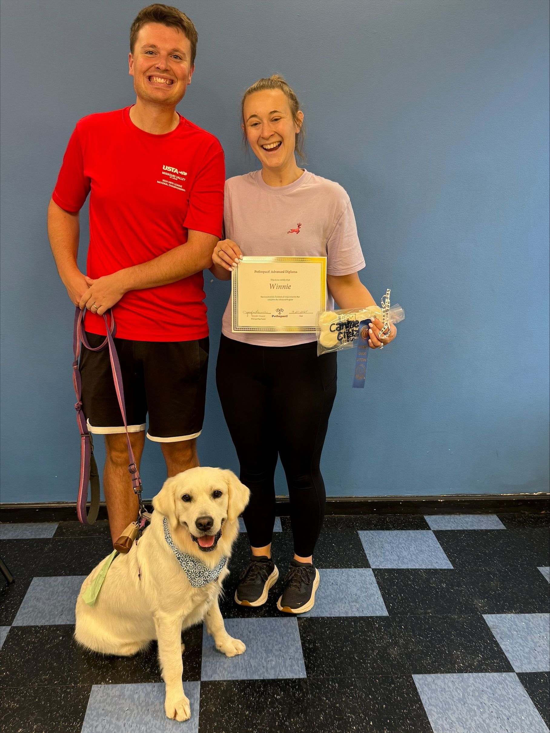 Man and woman with a golden lab, holding a certificate, in front of a blue wall.