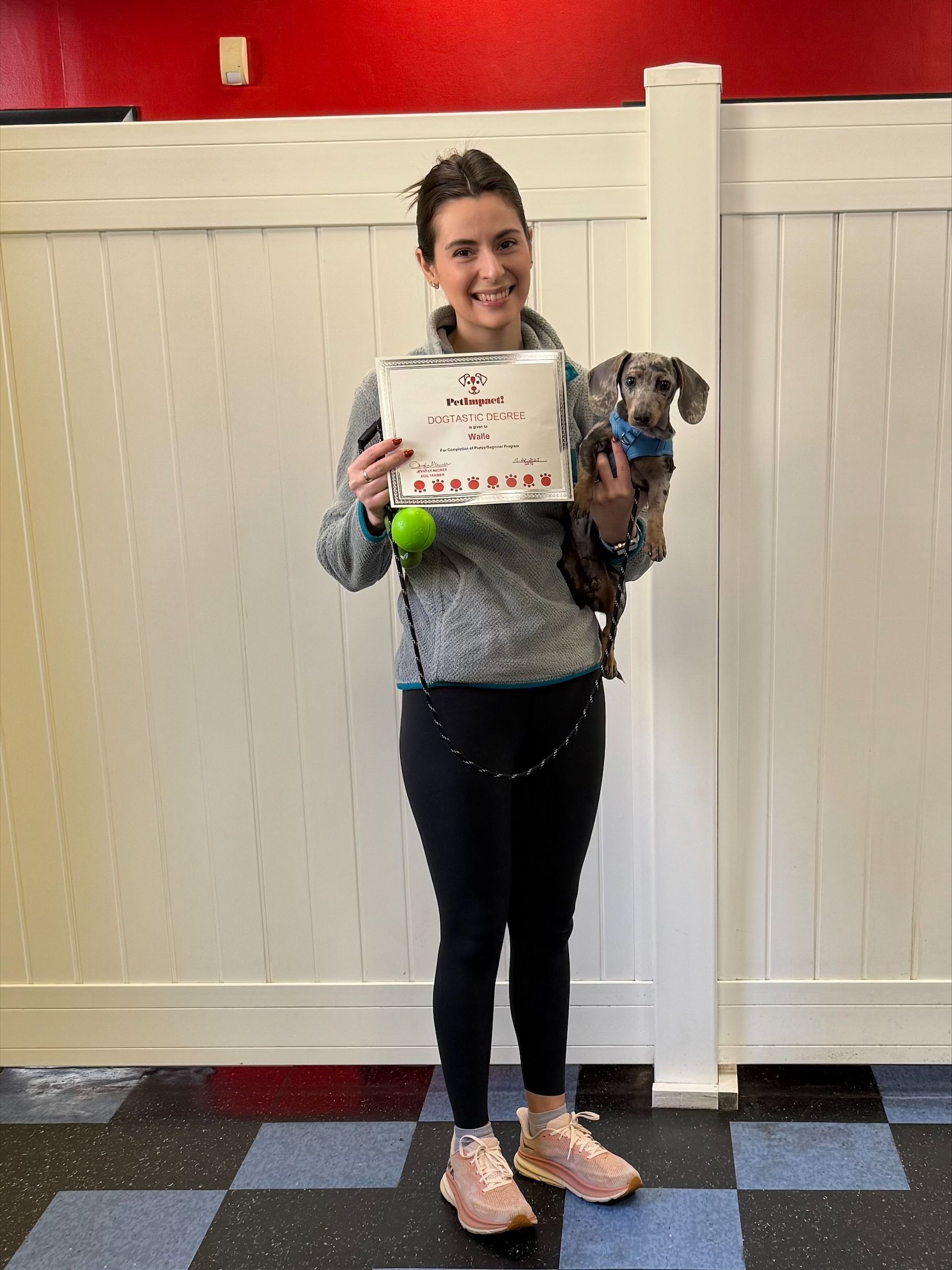 Woman with a dog holds a certificate, standing in a room with white paneling and a checkered floor.