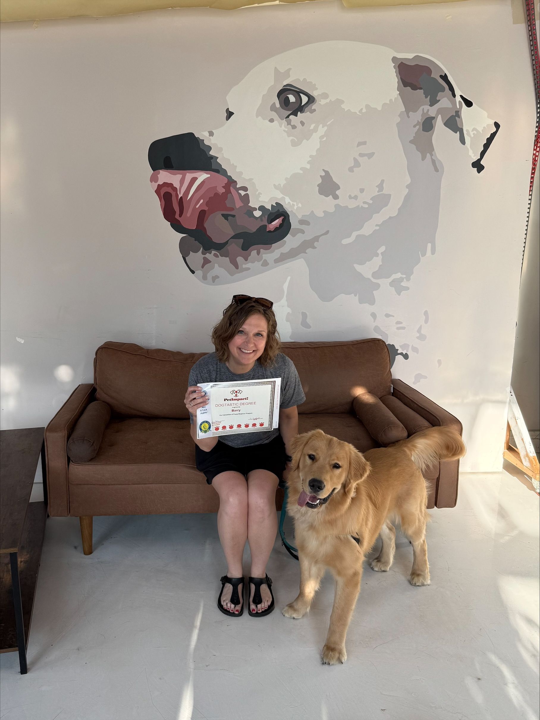 Woman and golden retriever pose with a certificate in front of a large dog painting, near a brown couch.