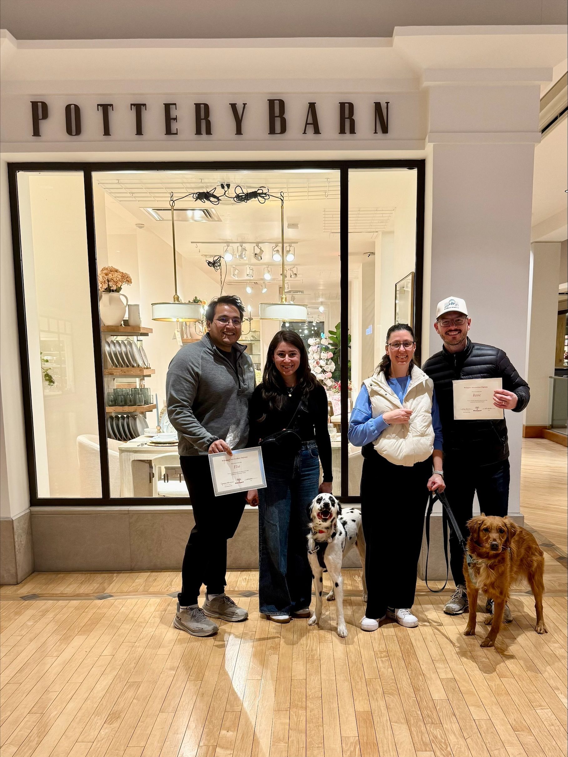 Four people and two dogs stand in front of Pottery Barn store. They hold papers, likely certificates.