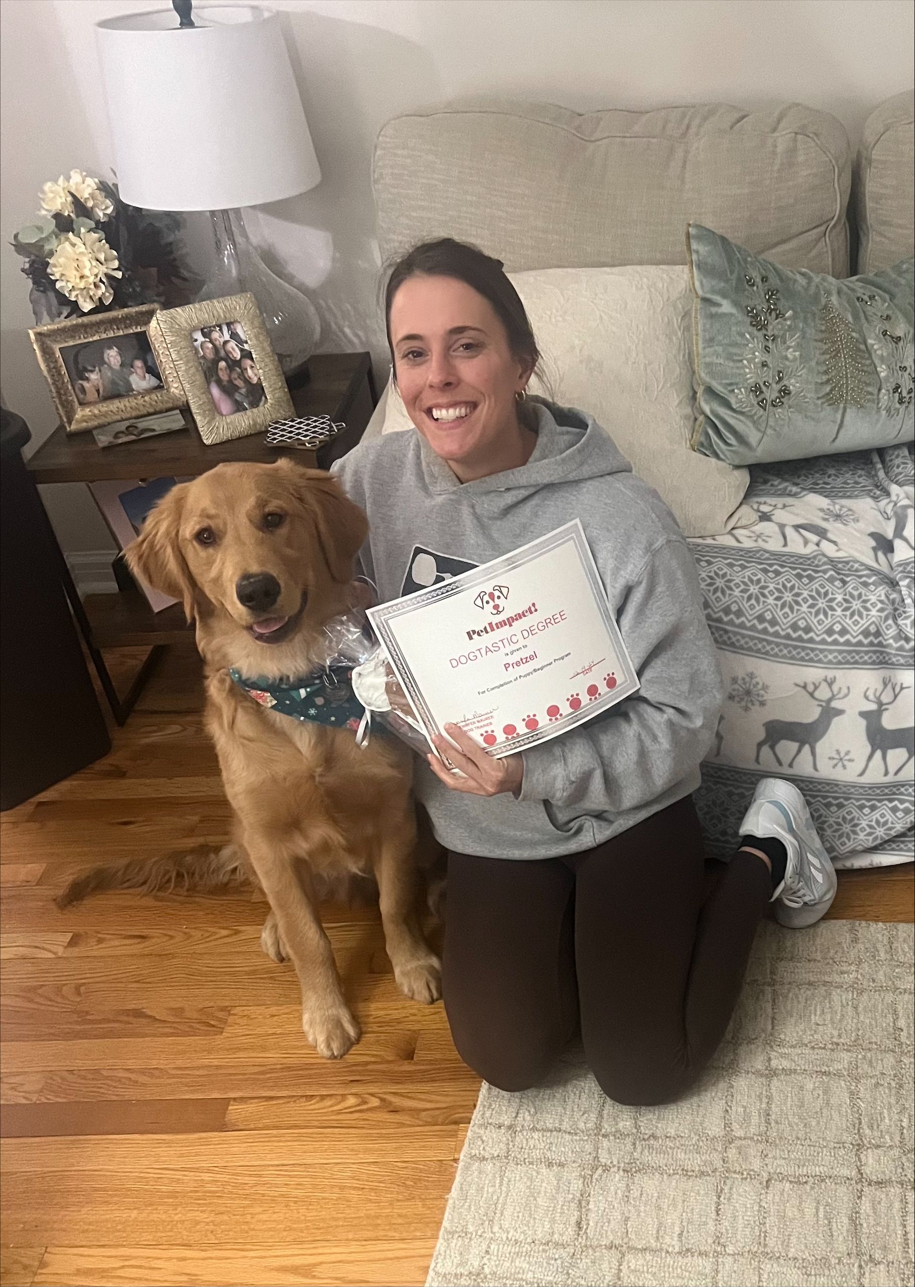 Woman kneeling with golden retriever, holding a certificate. Wooden floor, pillows, and a lamp in background.