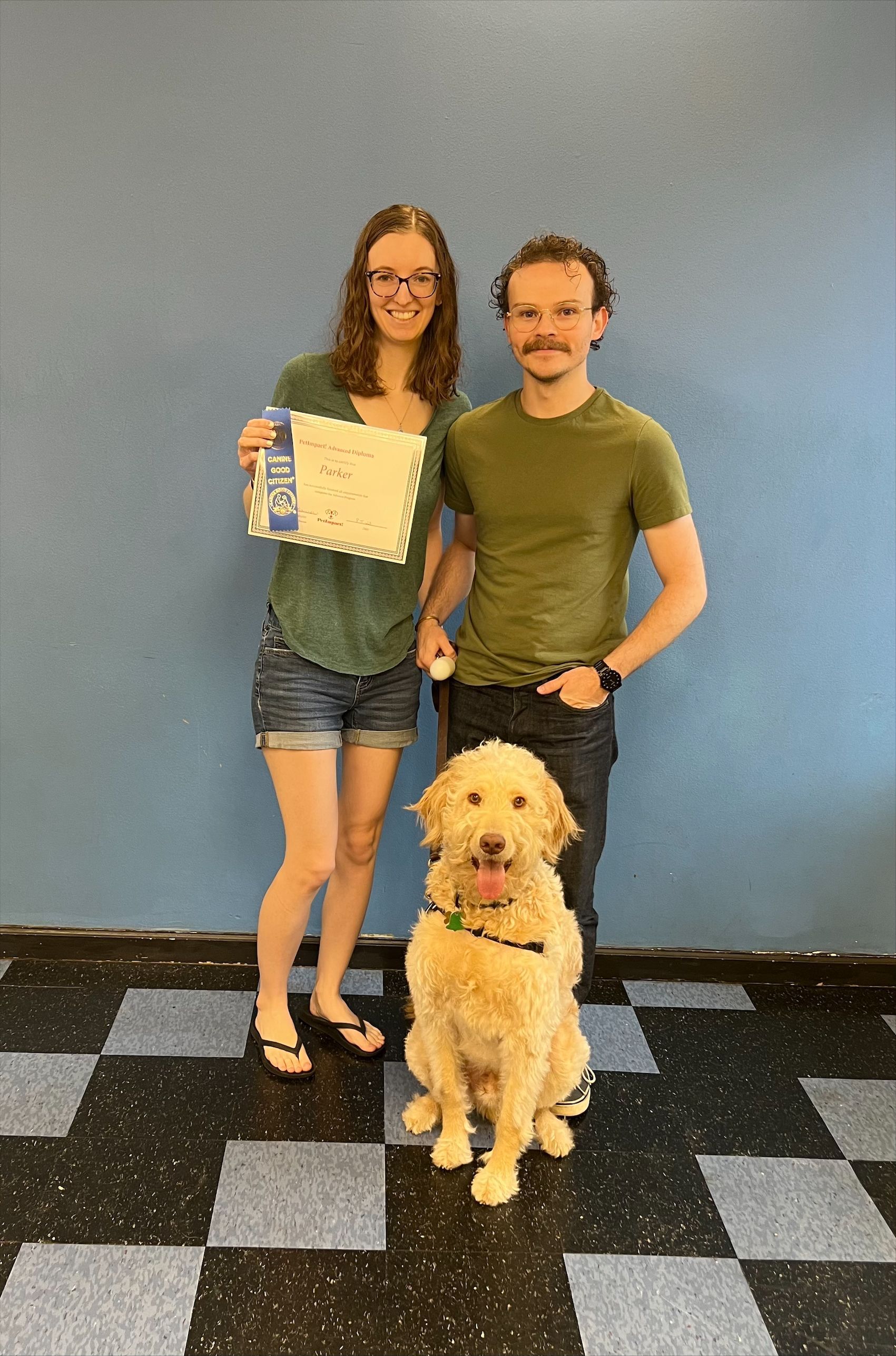 Couple and dog pose with an award in front of a blue wall and checkered floor.