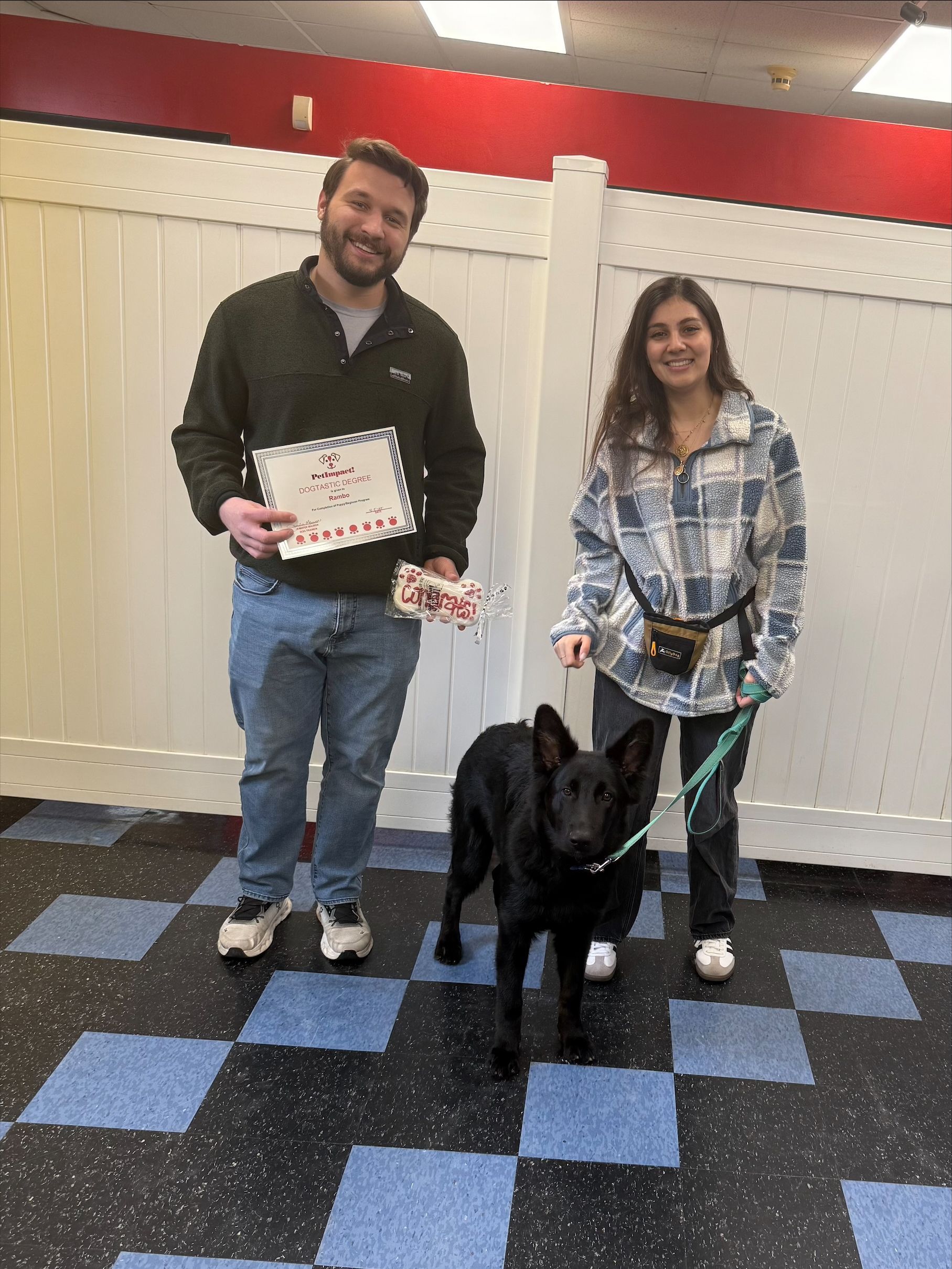 A man and a woman are standing next to a black dog on a leash.