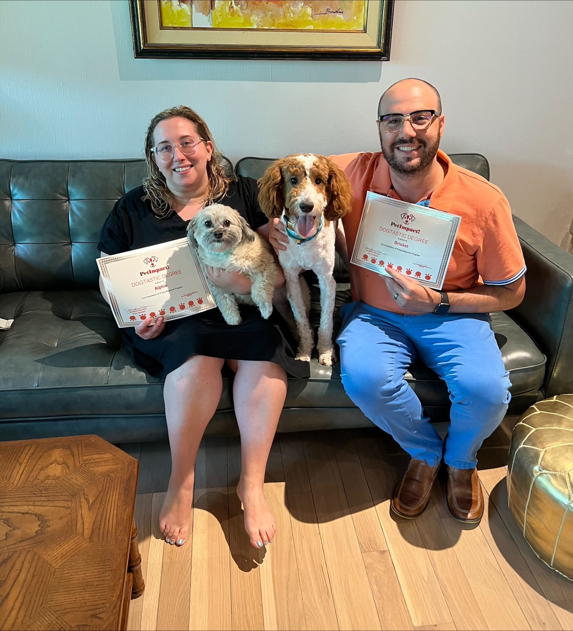 Couple and two dogs sitting on a couch, holding cards. One dog is held. Living room setting.