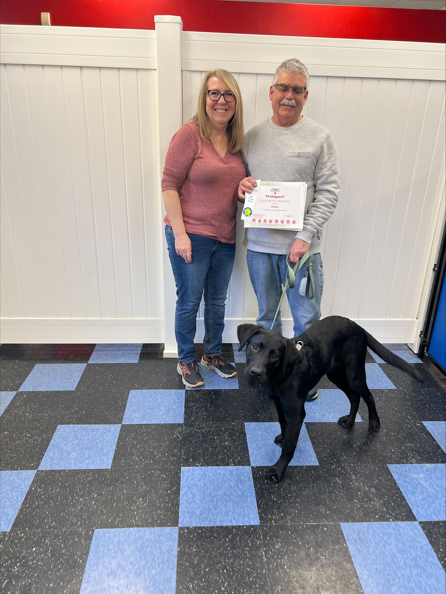 Woman, man, and black dog pose with a certificate in a room with checkered floor and white walls.