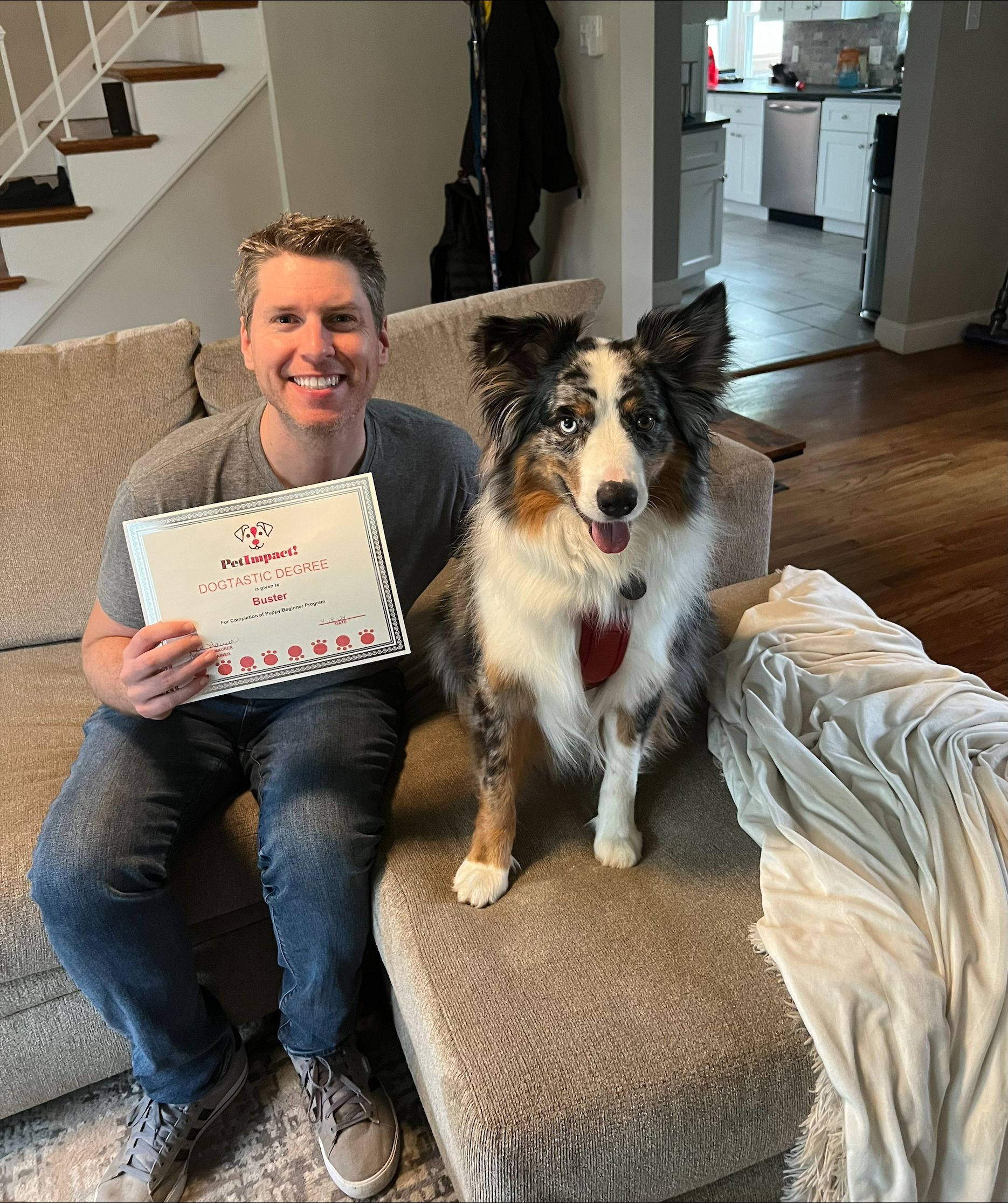Man and dog on couch. Man holds a card; the dog wears a heart collar. They are indoors.