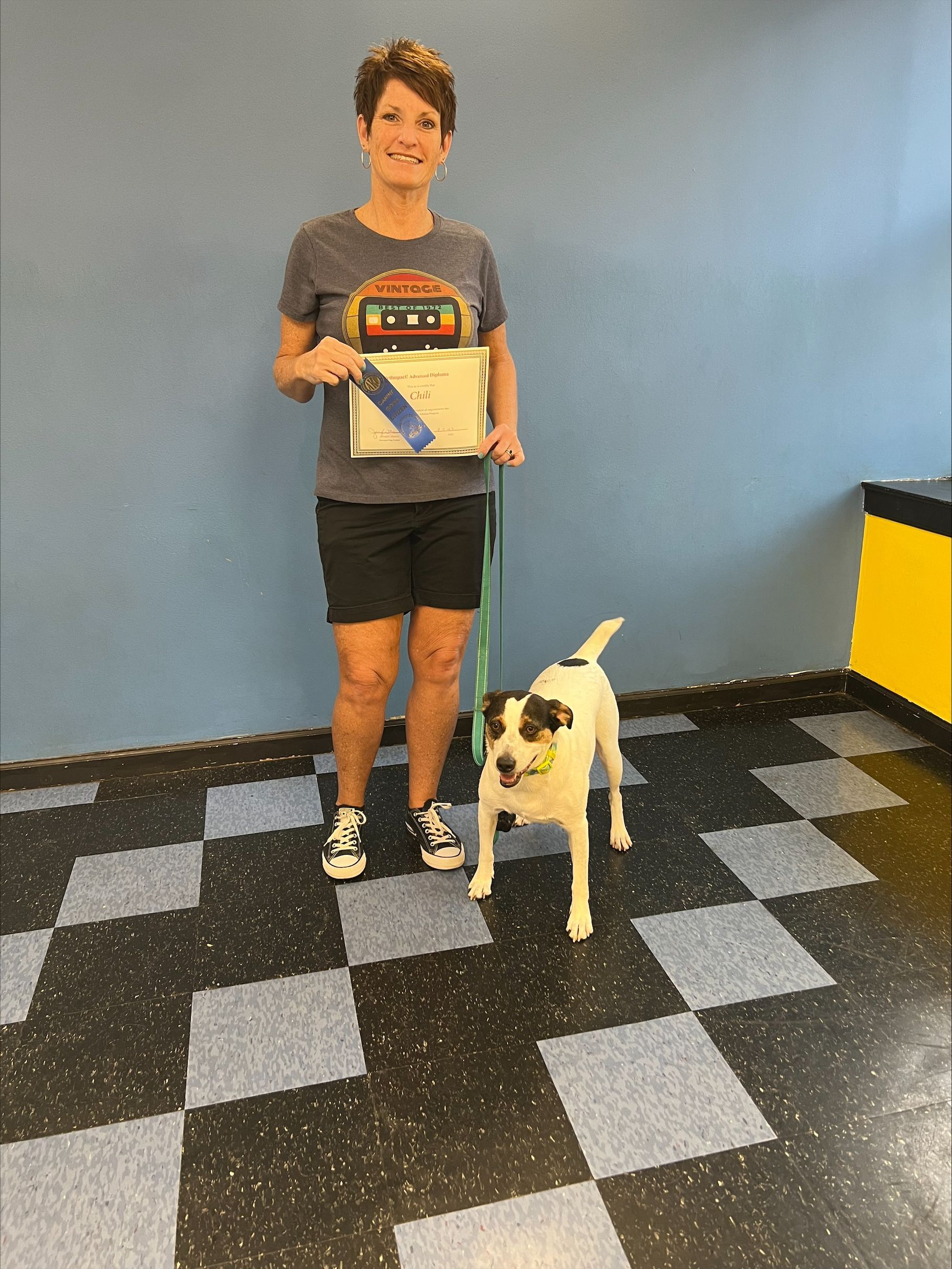 Woman and dog stand proudly next to each other. The woman is holding a certificate with a blue ribbon in a room with checkered floors.