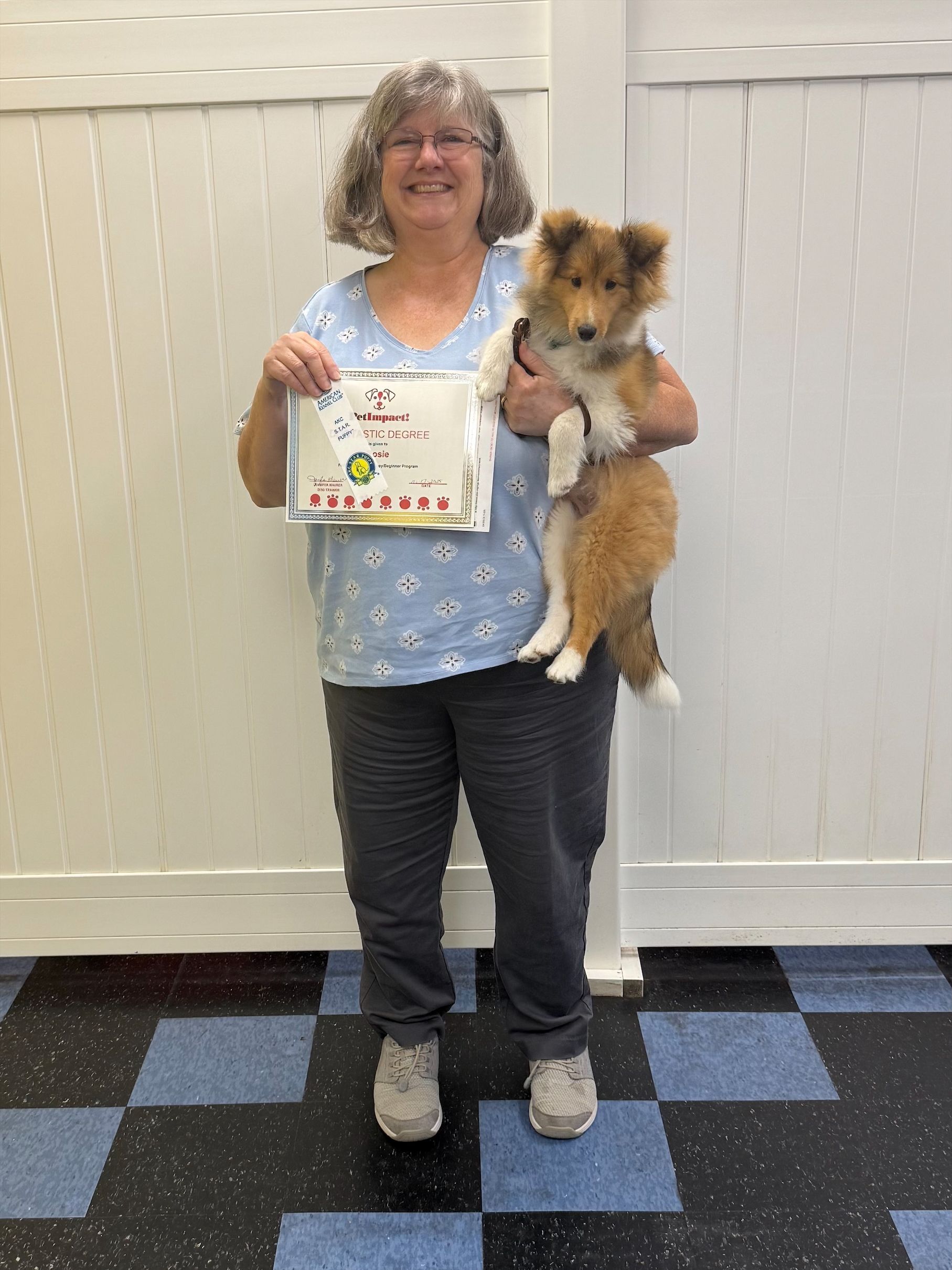 Woman holding Sheltie dog and a framed drawing; standing in front of a white wall, checkered floor.