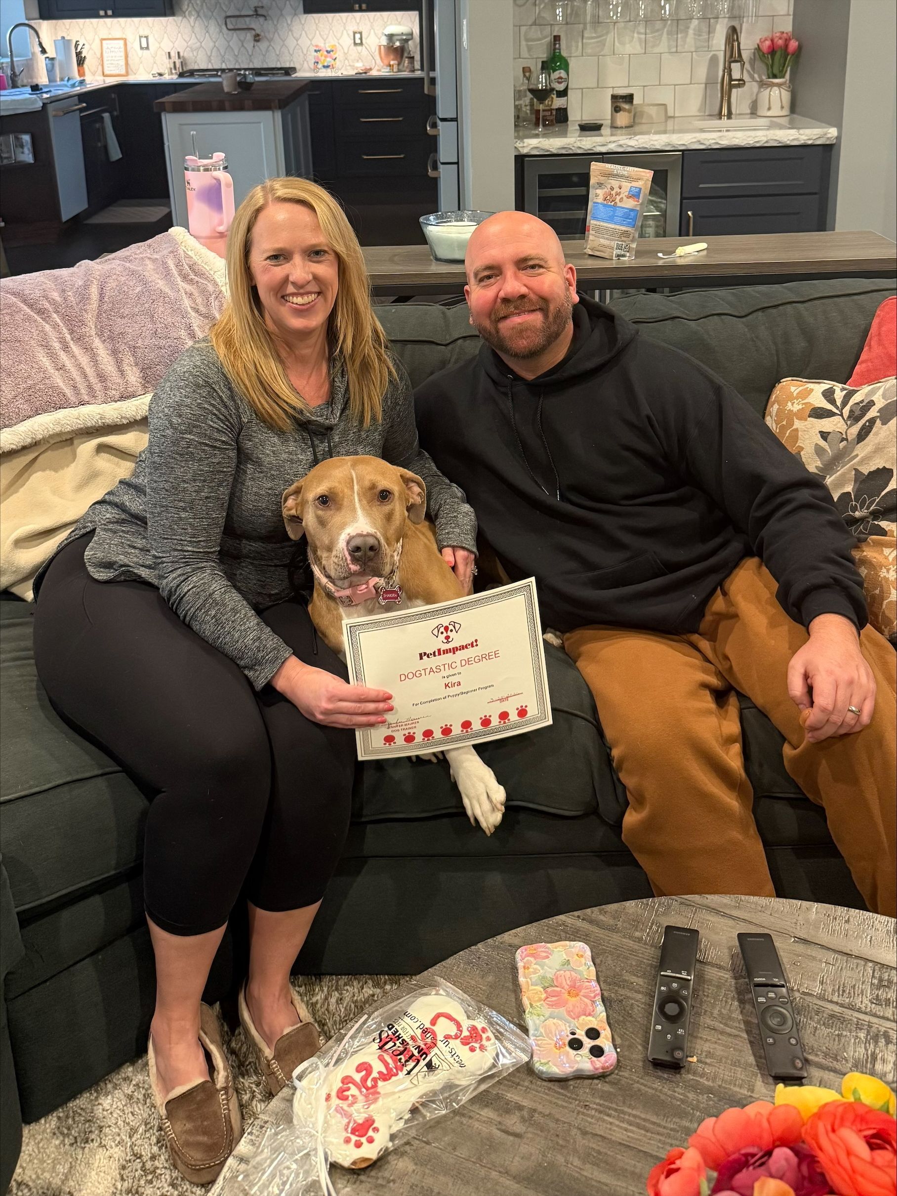 A couple sits on a dark sofa with their dog holding a sign, with celebratory treats on the coffee table in front of them.
