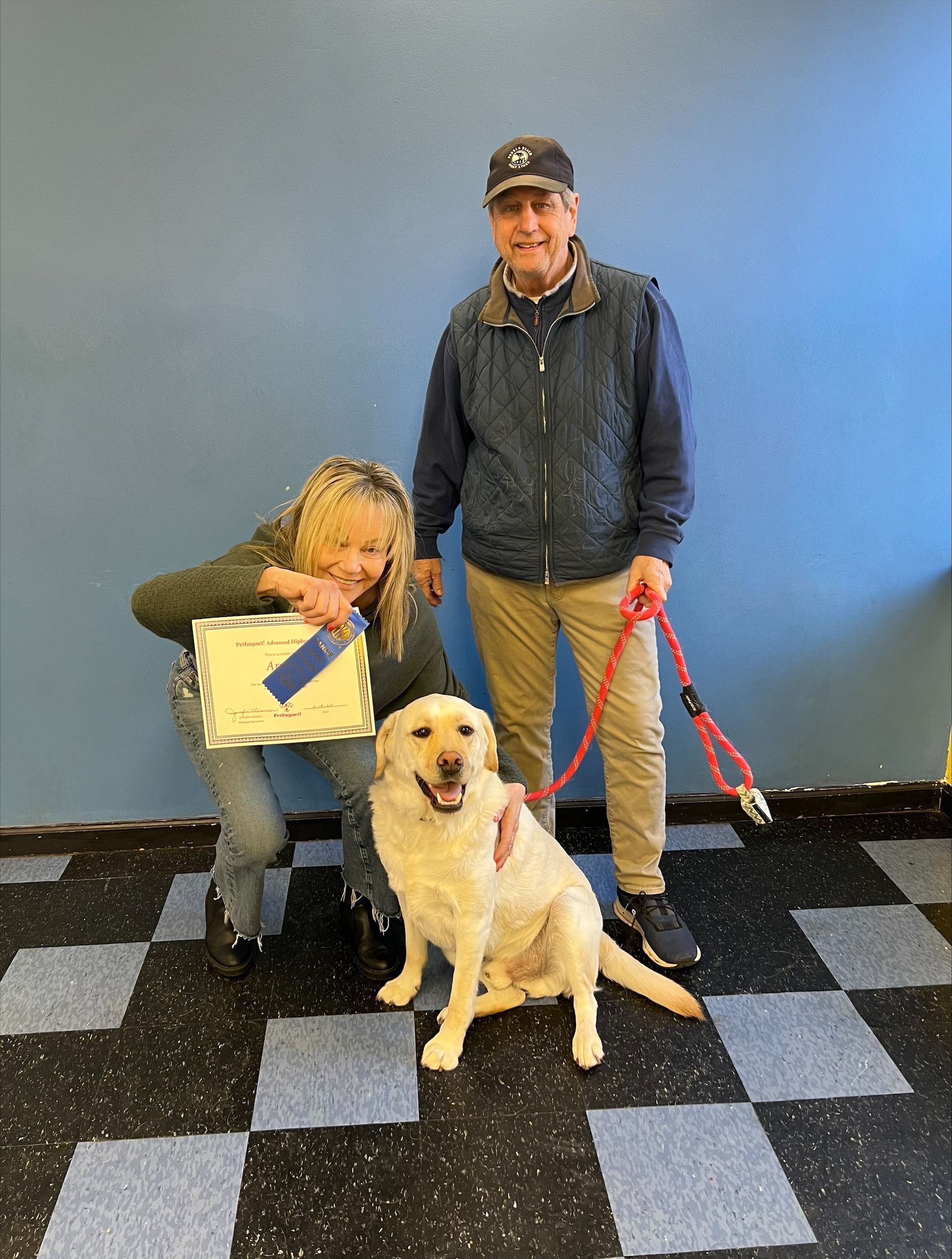 People with a yellow Labrador, the woman holds a certificate, and a man holds the leash; they stand in front of a blue wall.