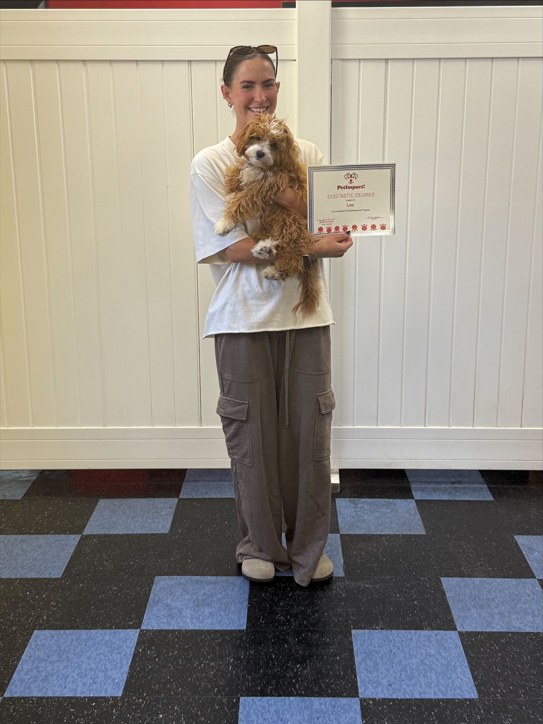 Woman holding a poodle, showing a certificate; indoors with checkered floor and white walls.
