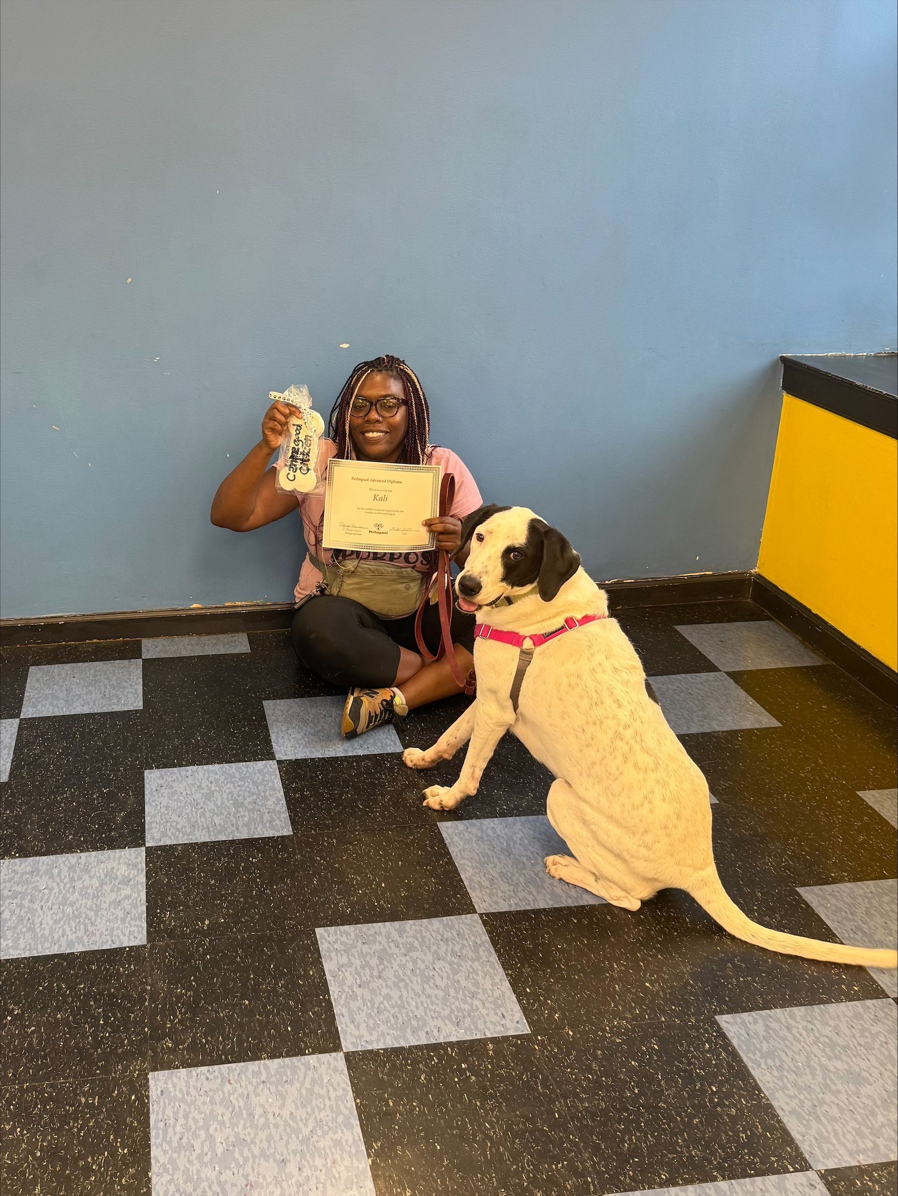 Woman and dog seated against blue wall. Woman smiles, holding certificate and gift; dog looks on. Black and white checkered floor.