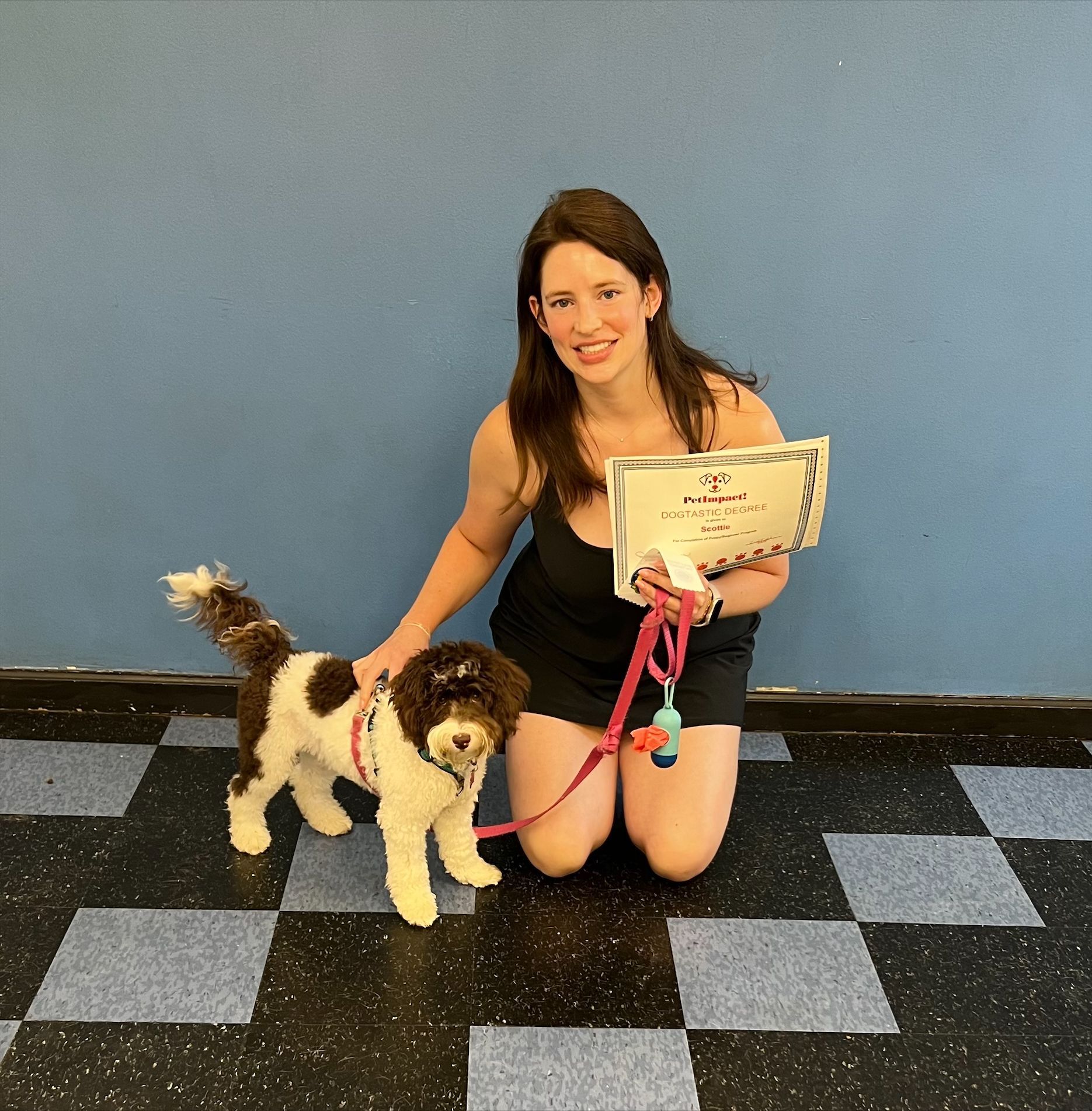 Woman kneeling with dog, holding certificate. Dog is white and brown, both smiling. Blue wall, checkered floor.