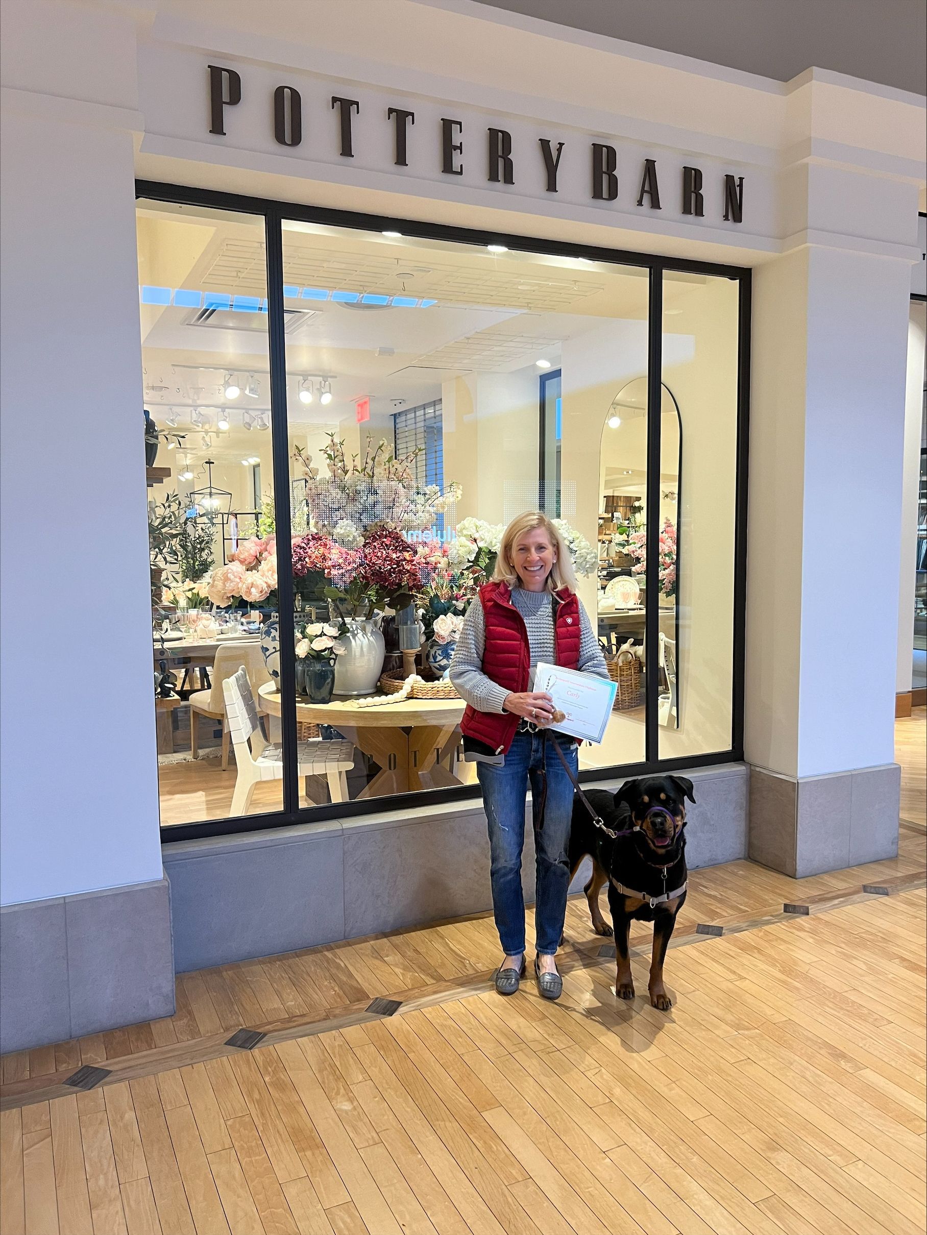 Woman and dog stand in front of Pottery Barn store. The woman wears a red vest and holds a white bag.