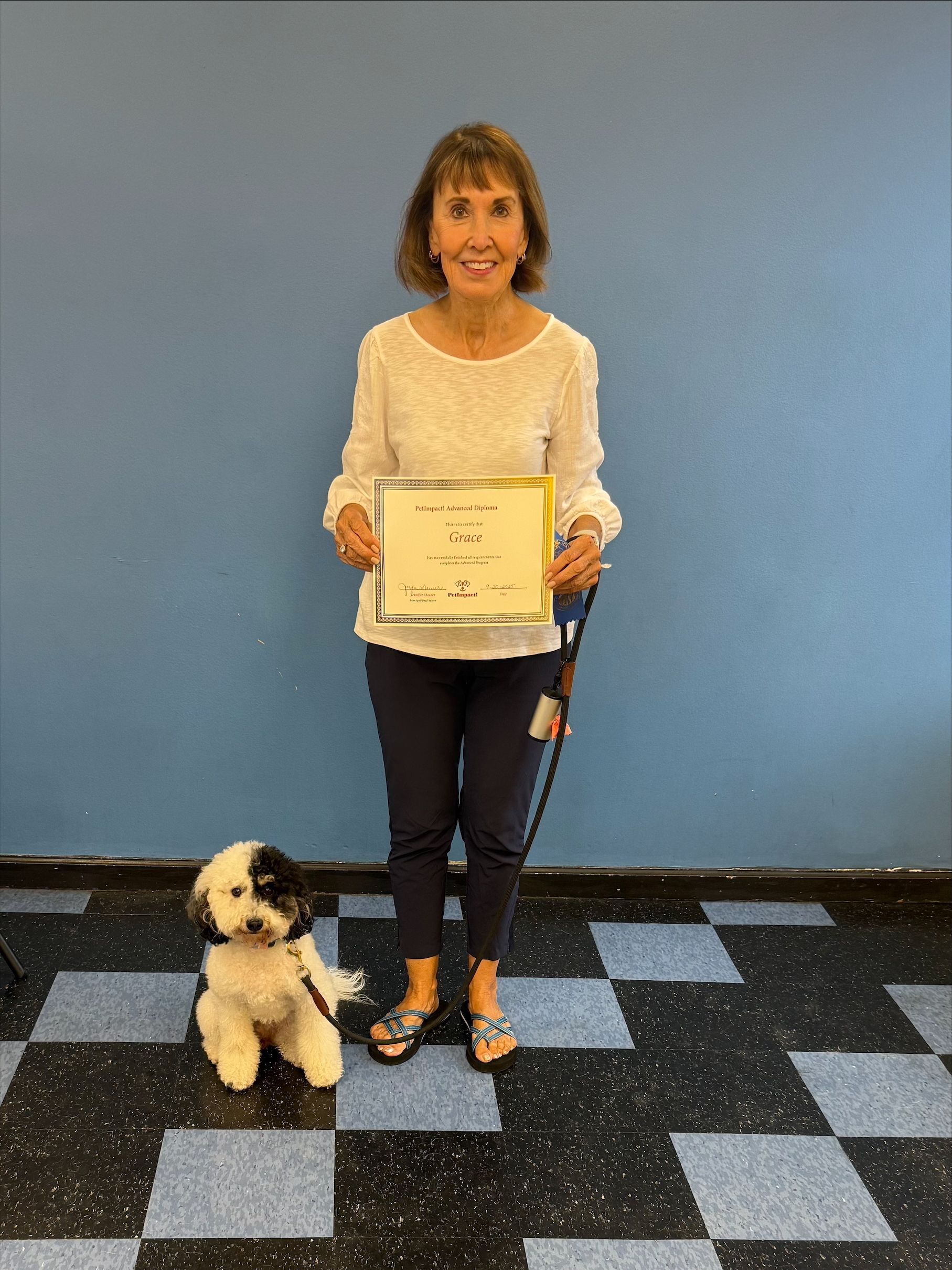 Woman with a certificate and dog; they're standing in front of a blue wall with a checkered floor.