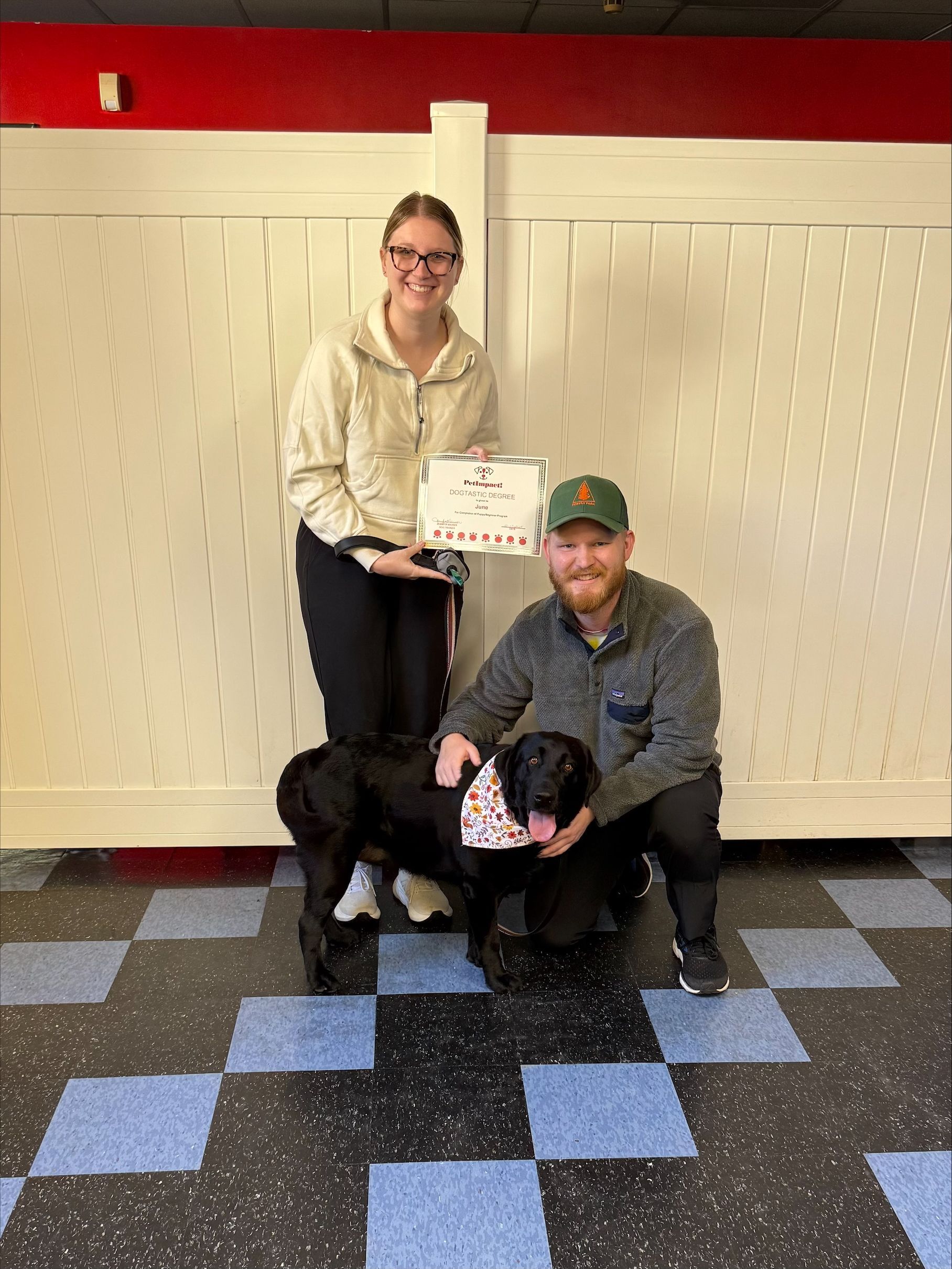 A woman and a man pose with a black dog, holding a certificate. They are in front of a white fence.