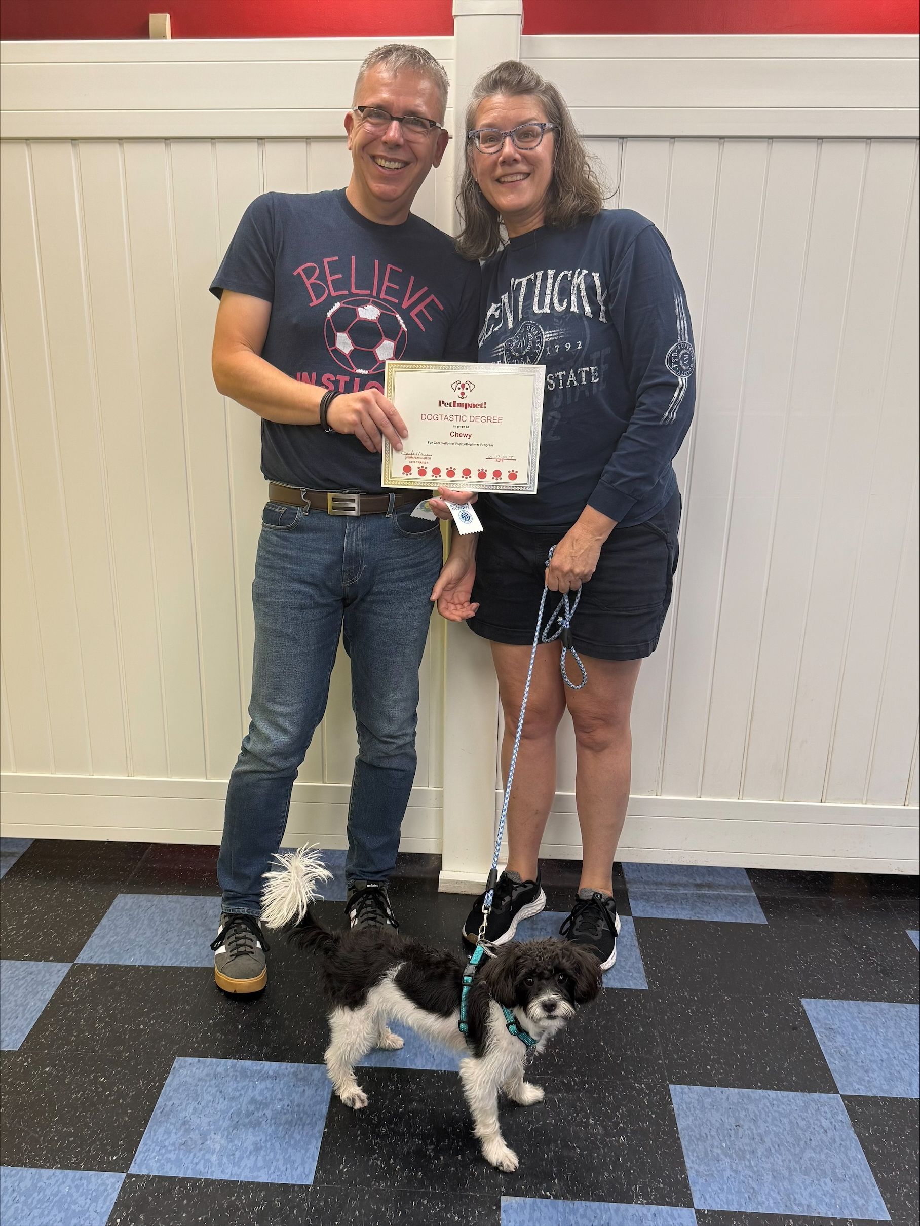 Two people and a small dog stand indoors, holding a certificate.