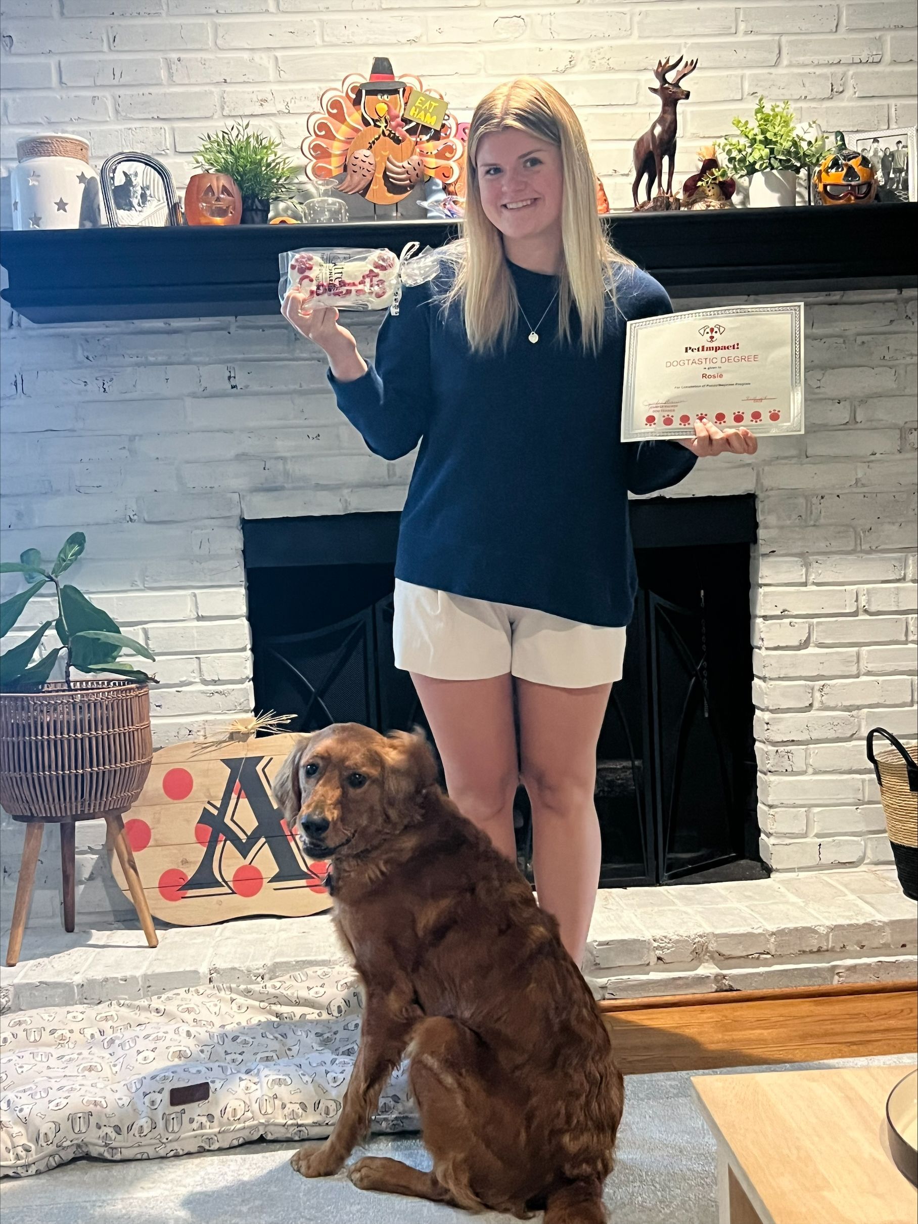A woman is standing next to a dog in a living room holding a pizza box.