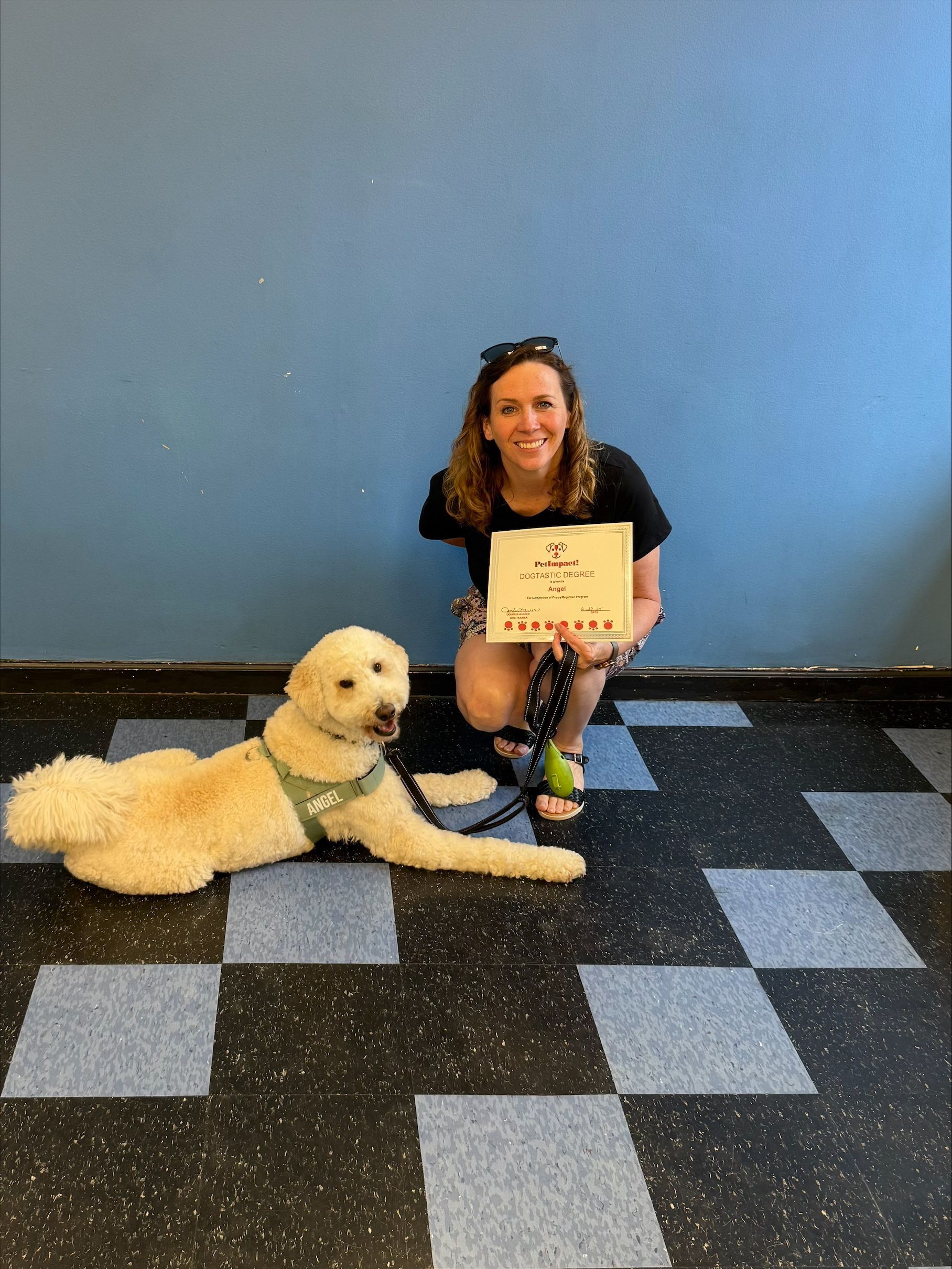 Woman smiles with dog, holding certificate in front of a blue wall and checkered floor.